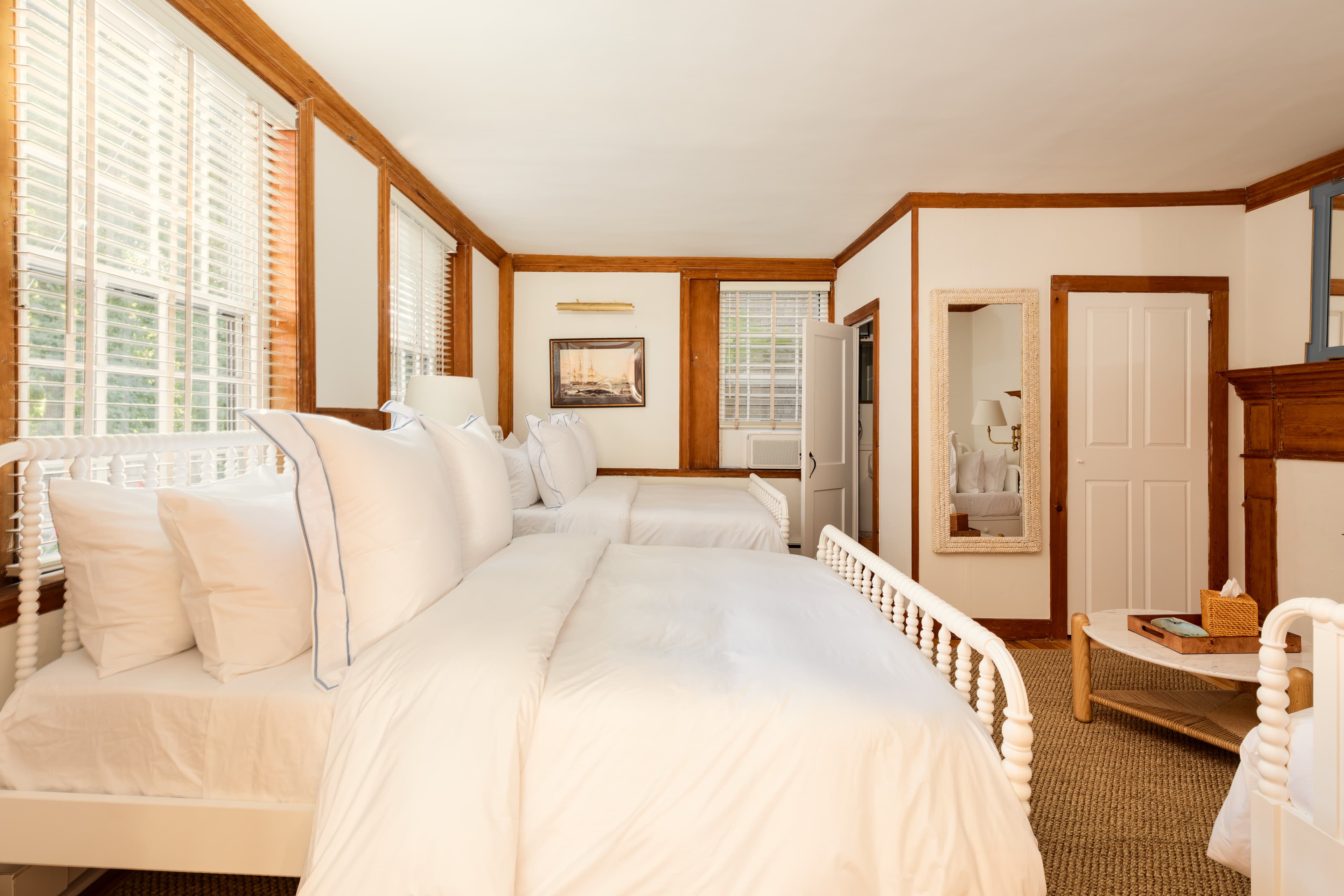 A bright, inviting bedroom featuring two white beds with crisp linens, wooden trim, and sunlit windows.