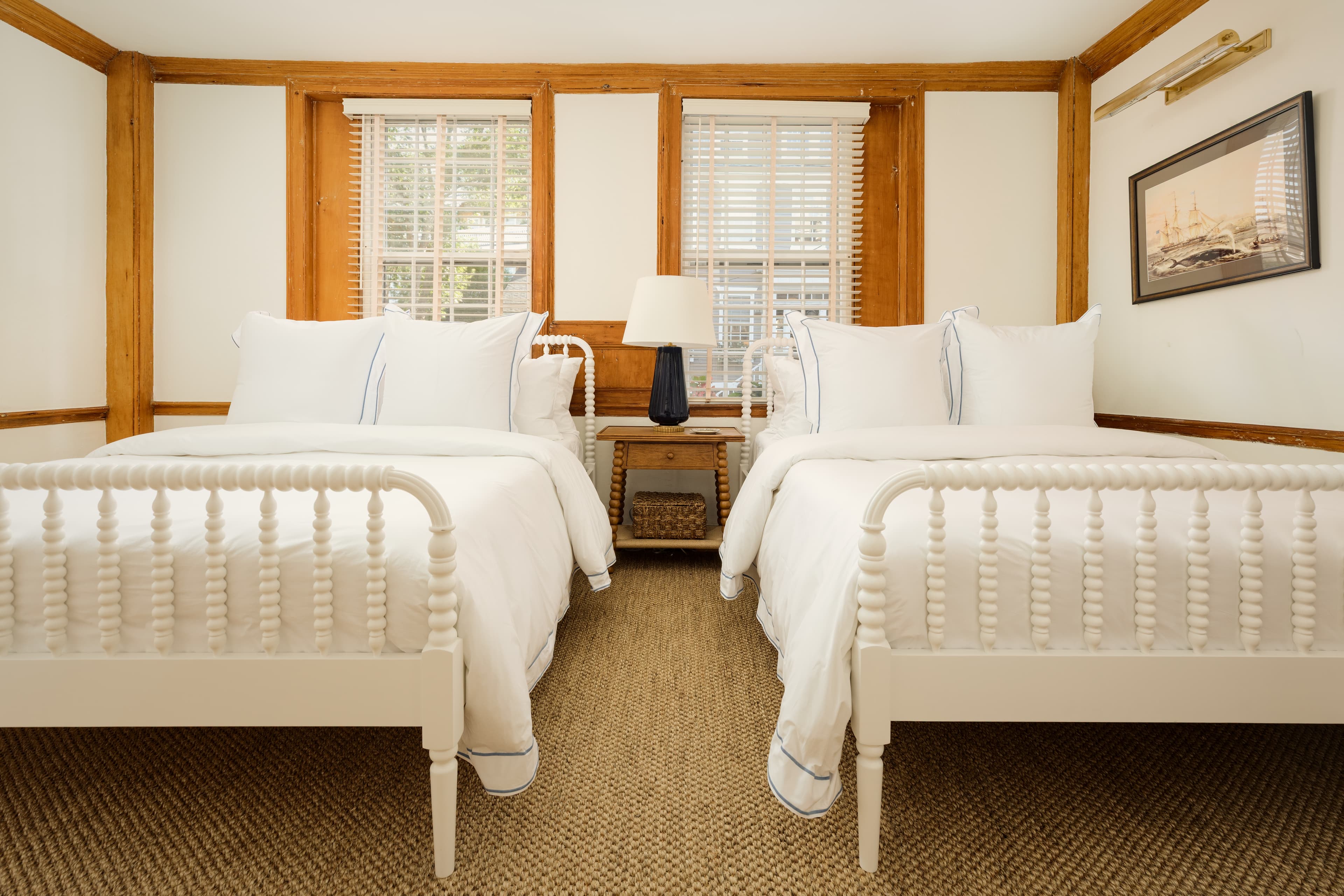 A charming bedroom with two white beds, light wood trim, and a nightstand with a lamp between them.