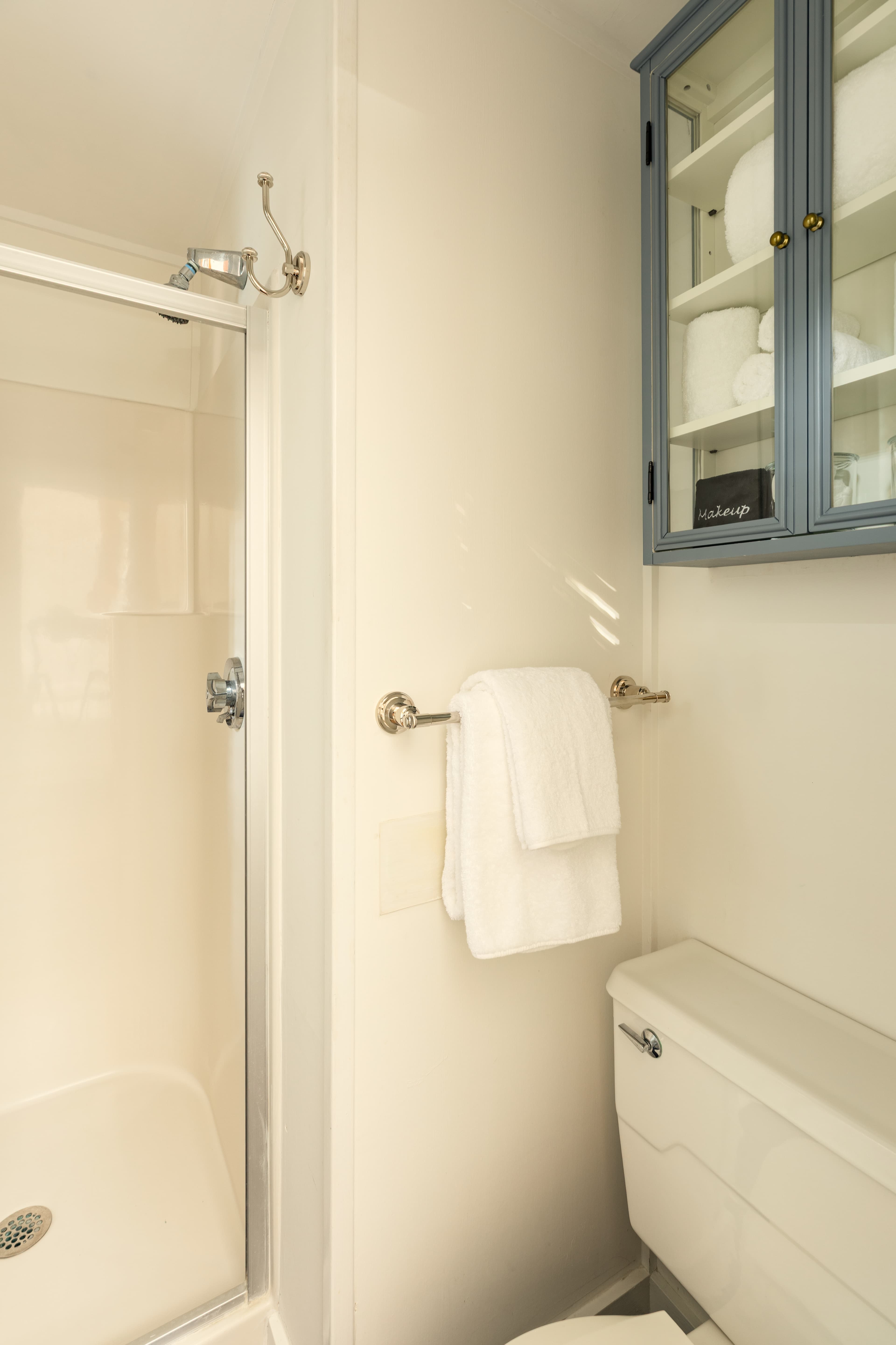 A compact, bright bathroom with a glass-enclosed shower, a white toilet, a towel bar with folded towels, and a blue wall cabinet.