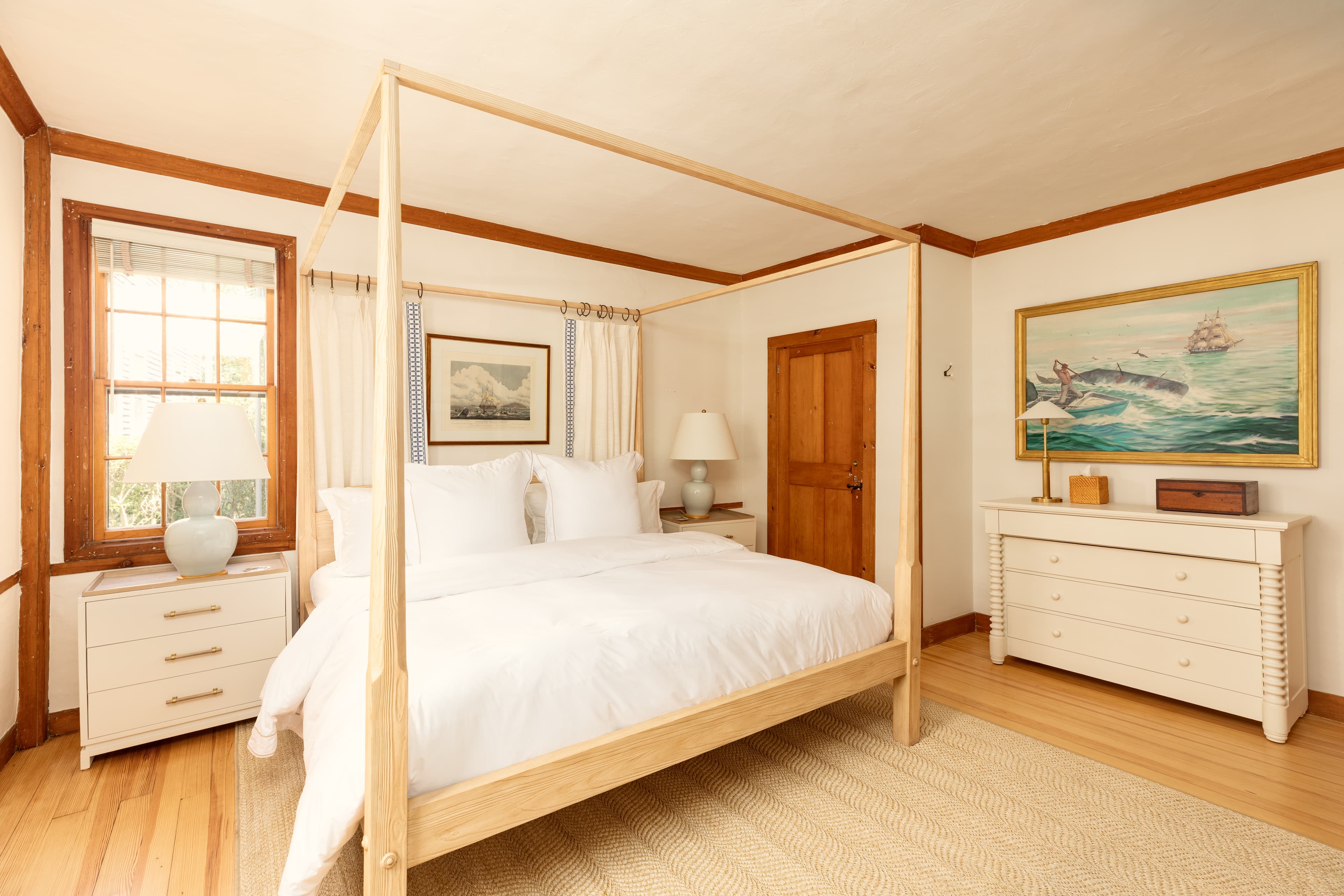 A spacious, sunlit bedroom featuring a light wood four-poster bed with white linens, matching dressers, and warm wooden trim.