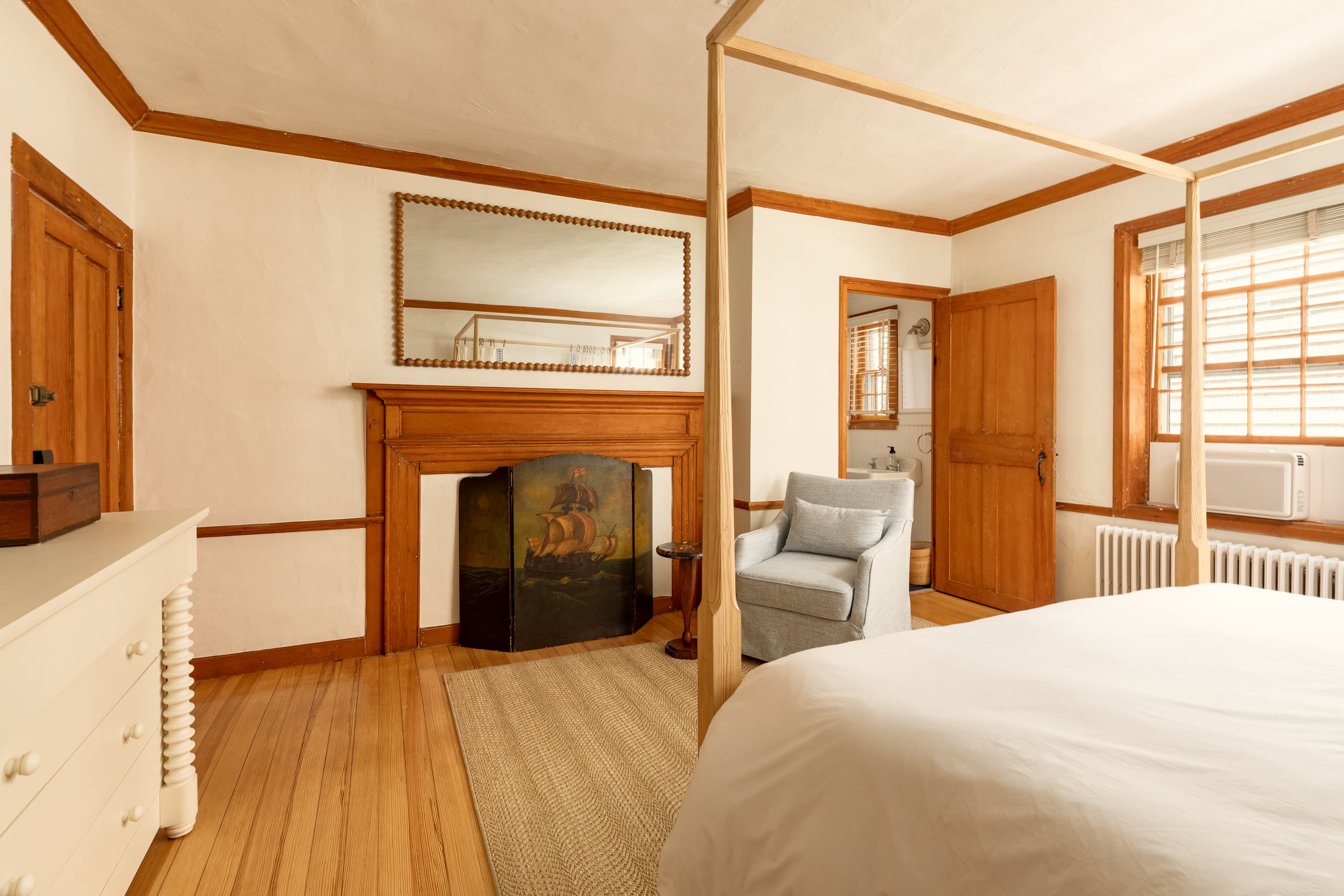 A spacious bedroom with a light wooden four-poster bed, a fireplace with a large mirror, a white dresser, and an armchair.