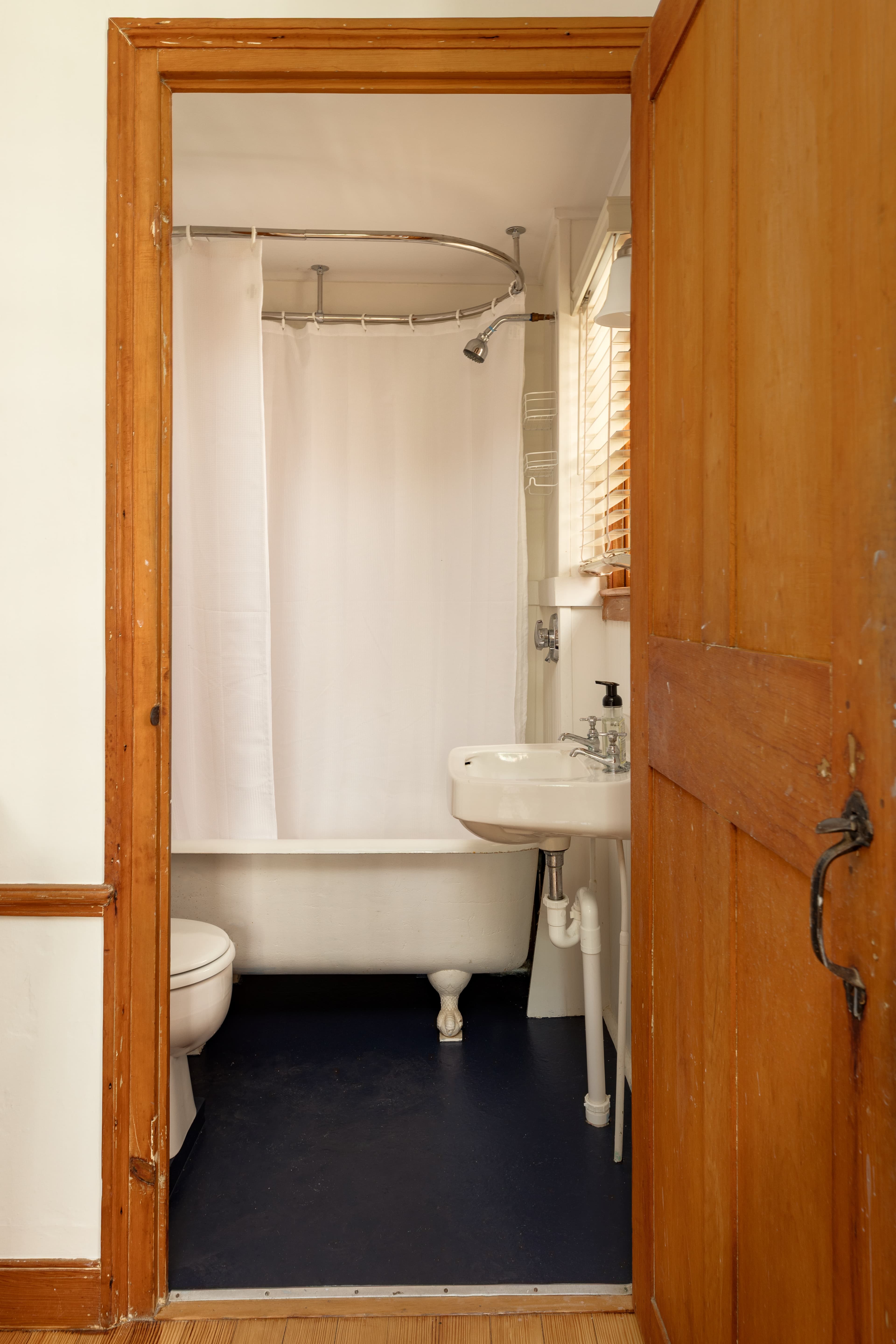 A small bathroom viewed through an open wooden doorway, featuring a claw-foot tub with a shower, a toilet, and a pedestal sink.