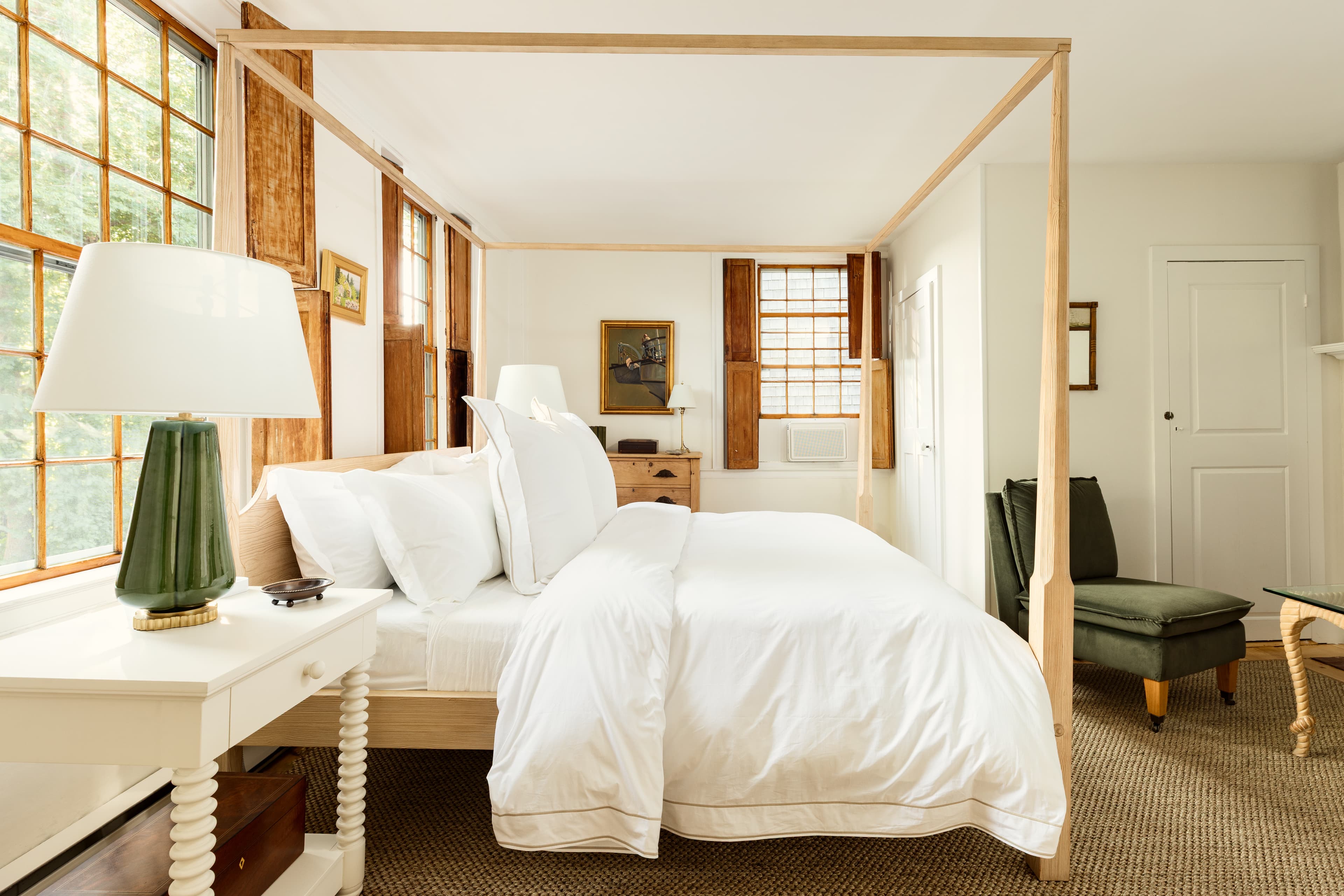 A serene bedroom featuring a light wood four-poster bed with white linens, a green lamp, and warm wooden accents, bathed in natural light.