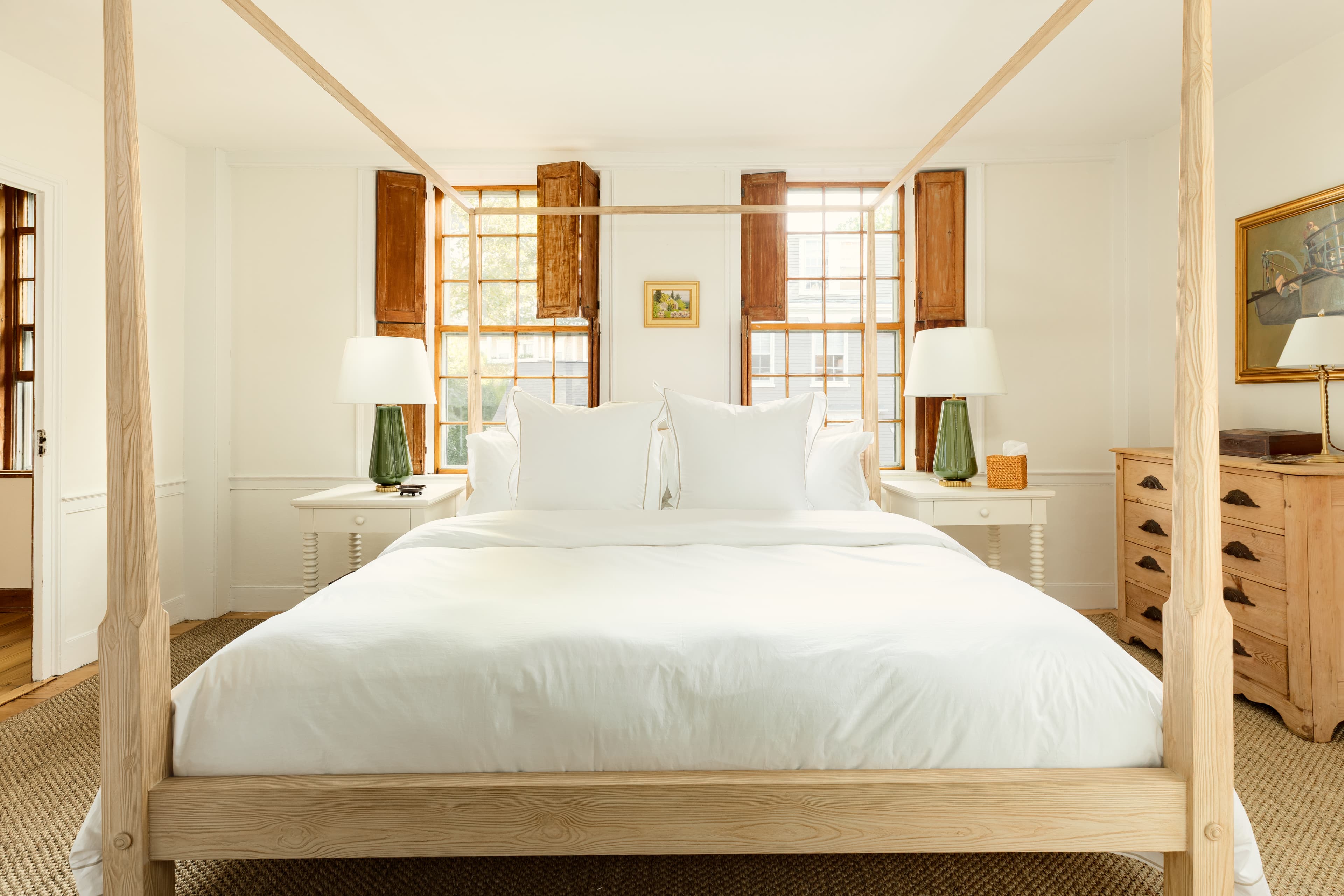 A sunlit bedroom with a light wood four-poster bed, crisp white linens, matching nightstands with green lamps, and warm wooden window shutters.