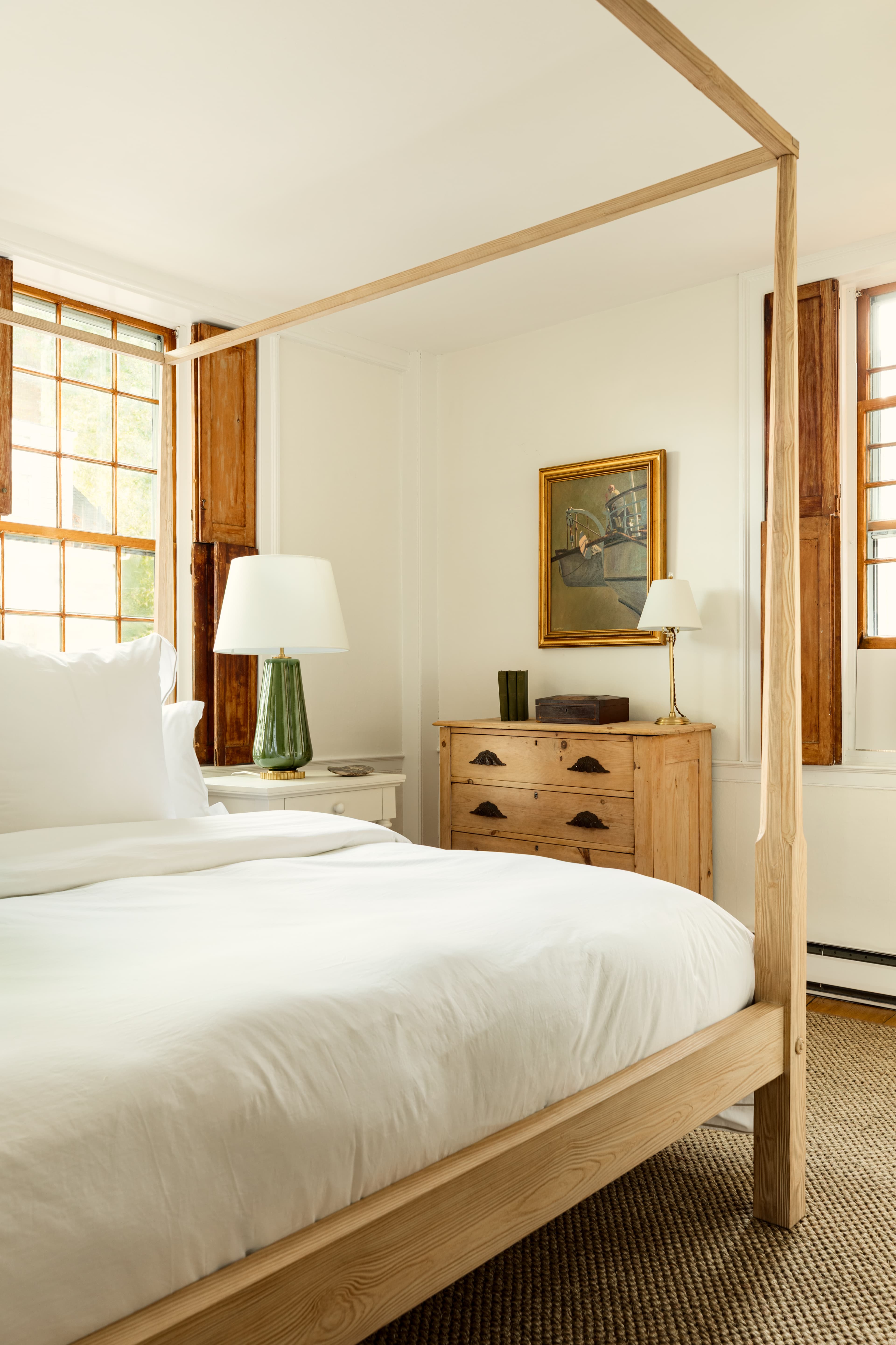 A cozy bedroom with a light wood four-poster bed, crisp white linens, a green lamp on a white nightstand, and wooden furniture, bathed in sunlight.