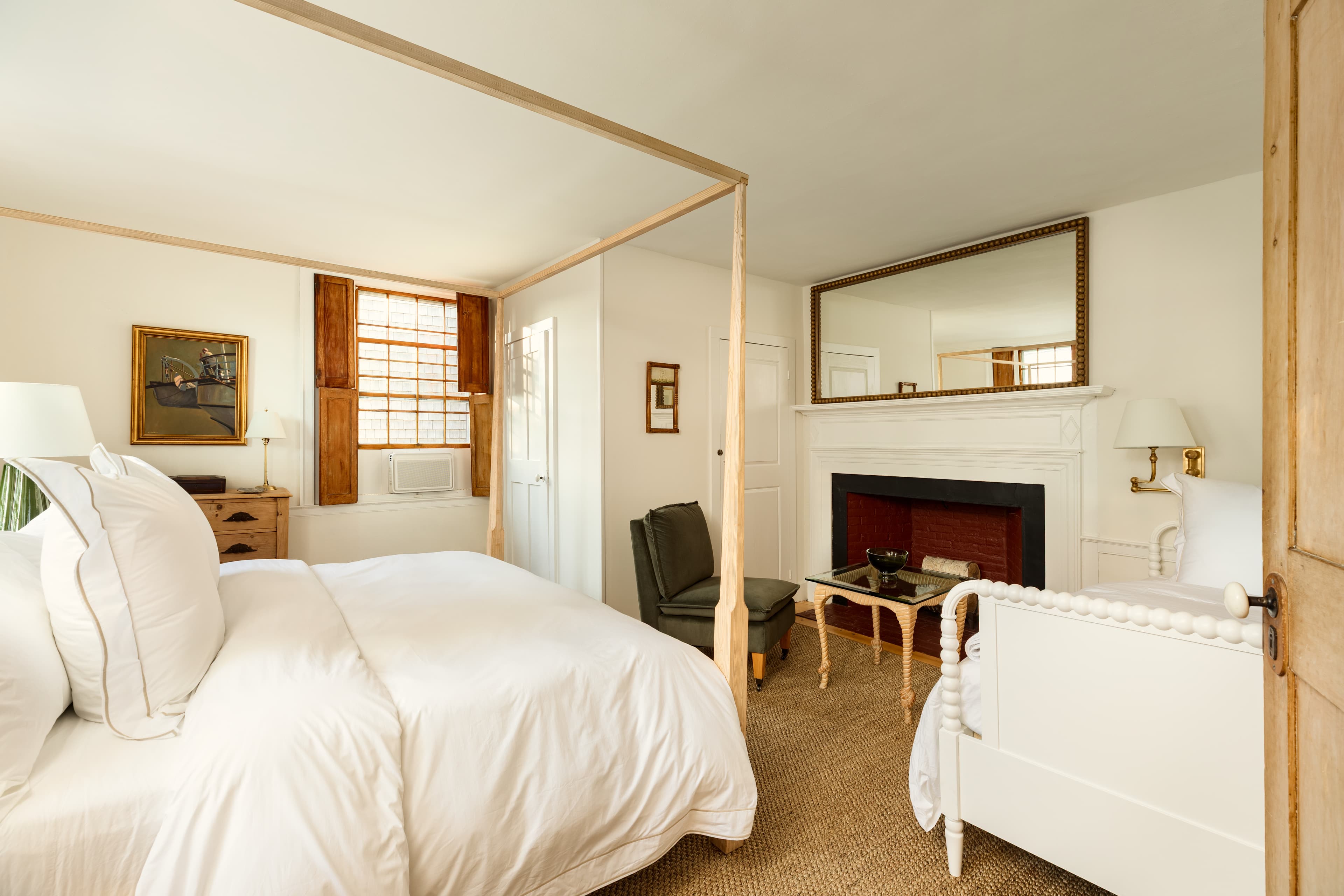 A large, sunlit bedroom with a light wood four-poster bed and a white daybed, featuring a fireplace with a mirror and warm wooden accents.