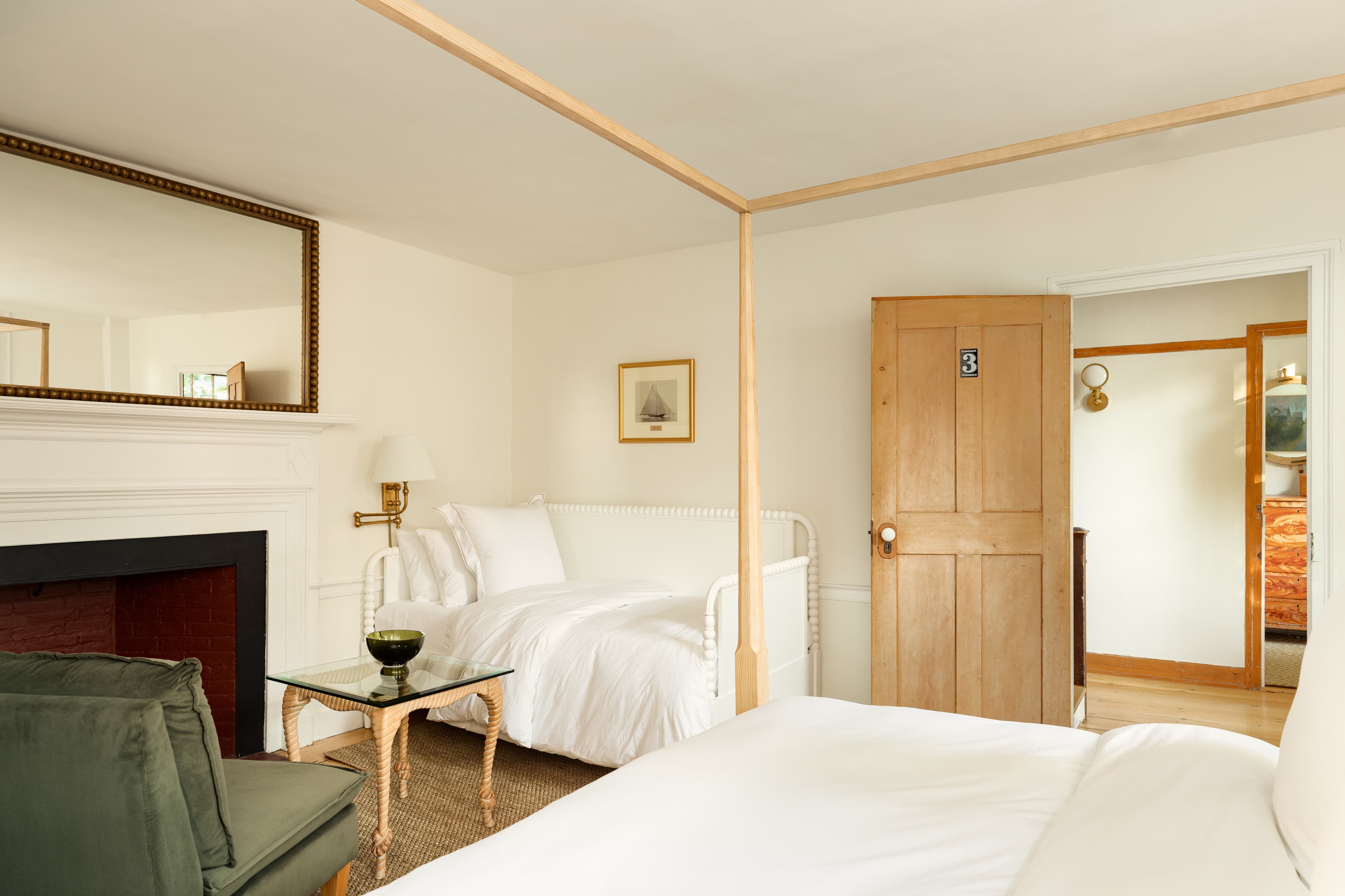 A cozy bedroom featuring a light wood four-poster bed and a white daybed, a fireplace with a large mirror, and a green armchair.