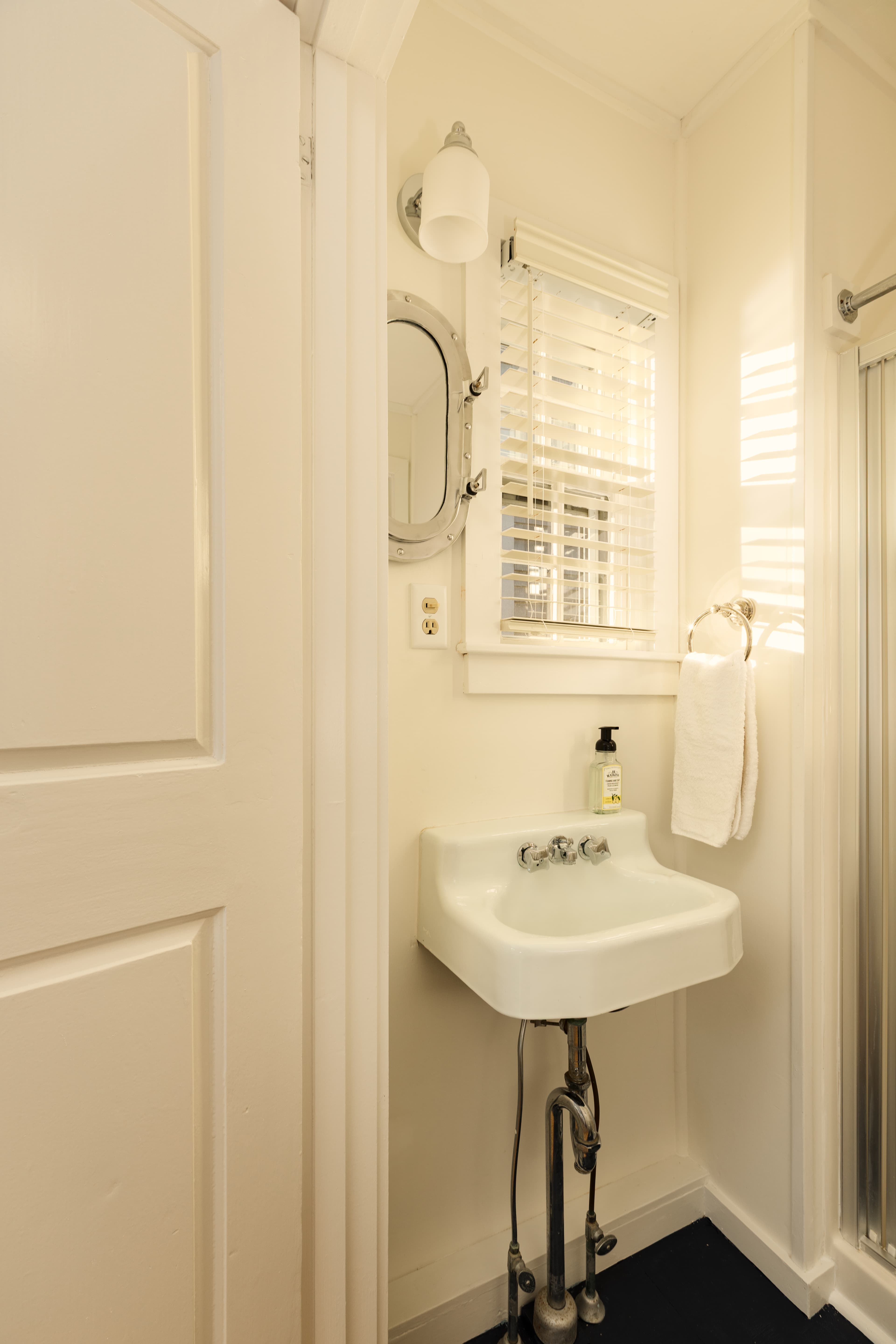 A small, brightly lit bathroom with a white pedestal sink, a window with blinds, a mirror, and a glass-enclosed shower on the right.
