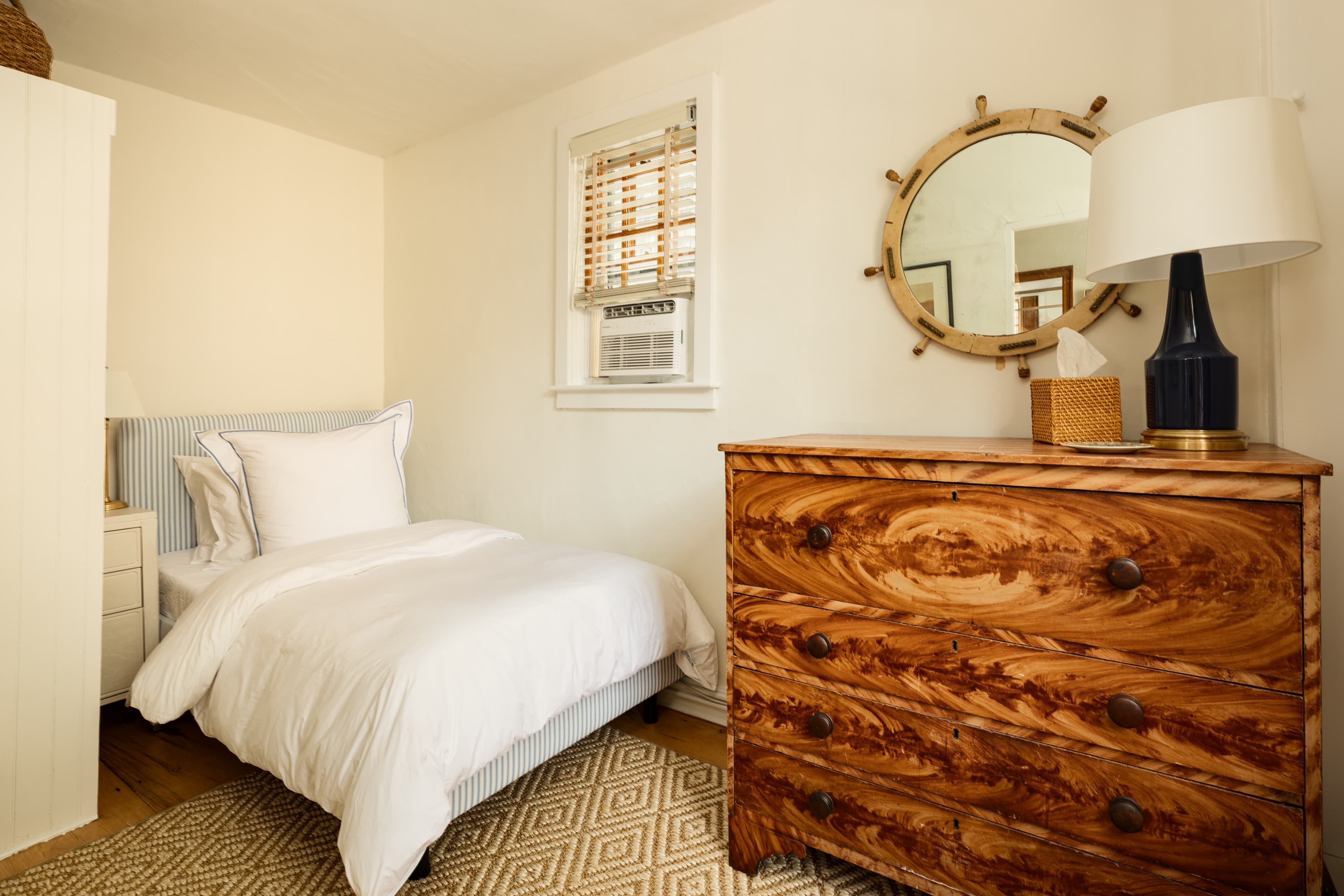A charming bedroom featuring a single white bed, a unique wooden dresser with a nautical-themed mirror, and a window with blinds.