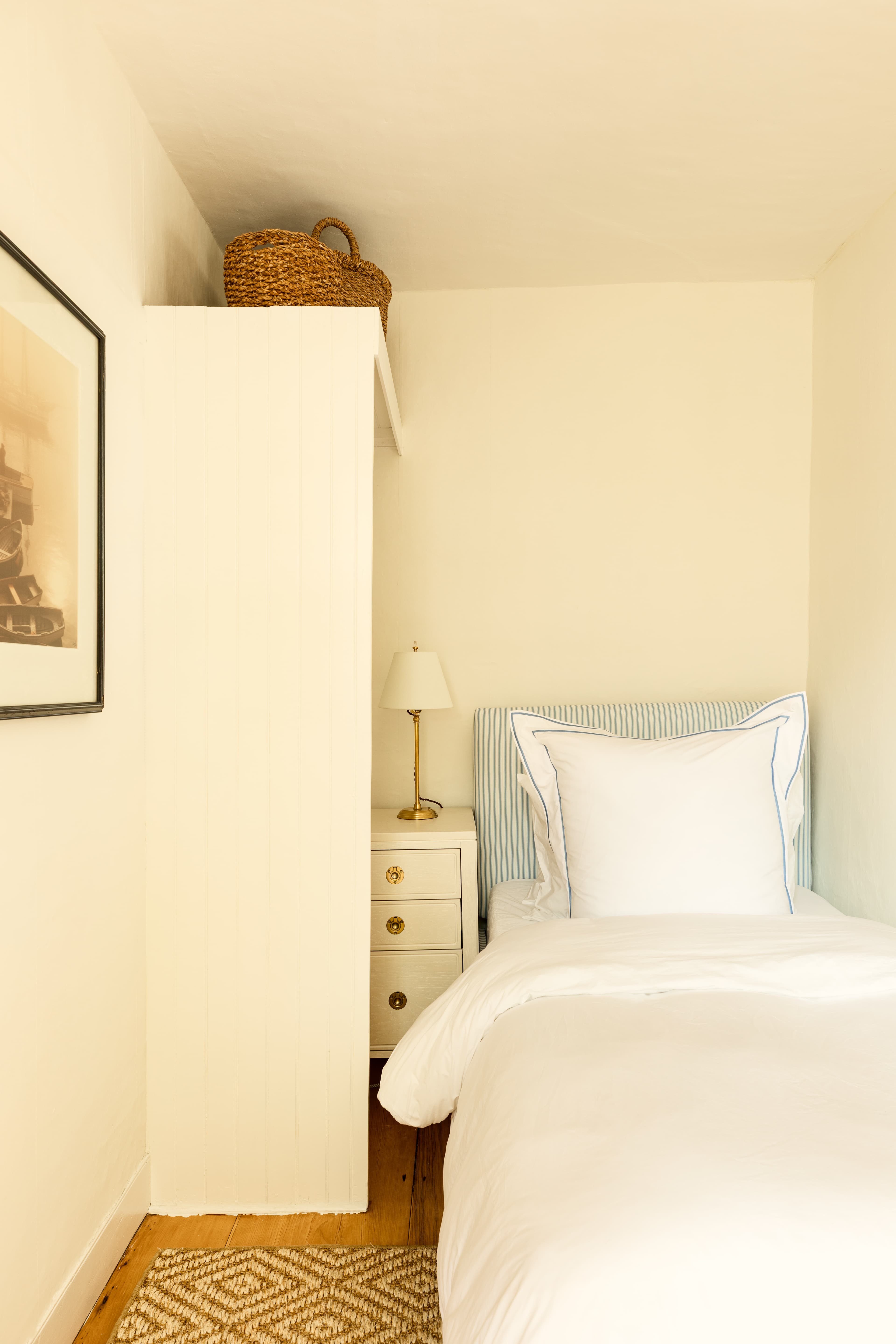 A small, cozy bedroom featuring a single bed with white linens, a white wardrobe, and a small nightstand with a lamp.