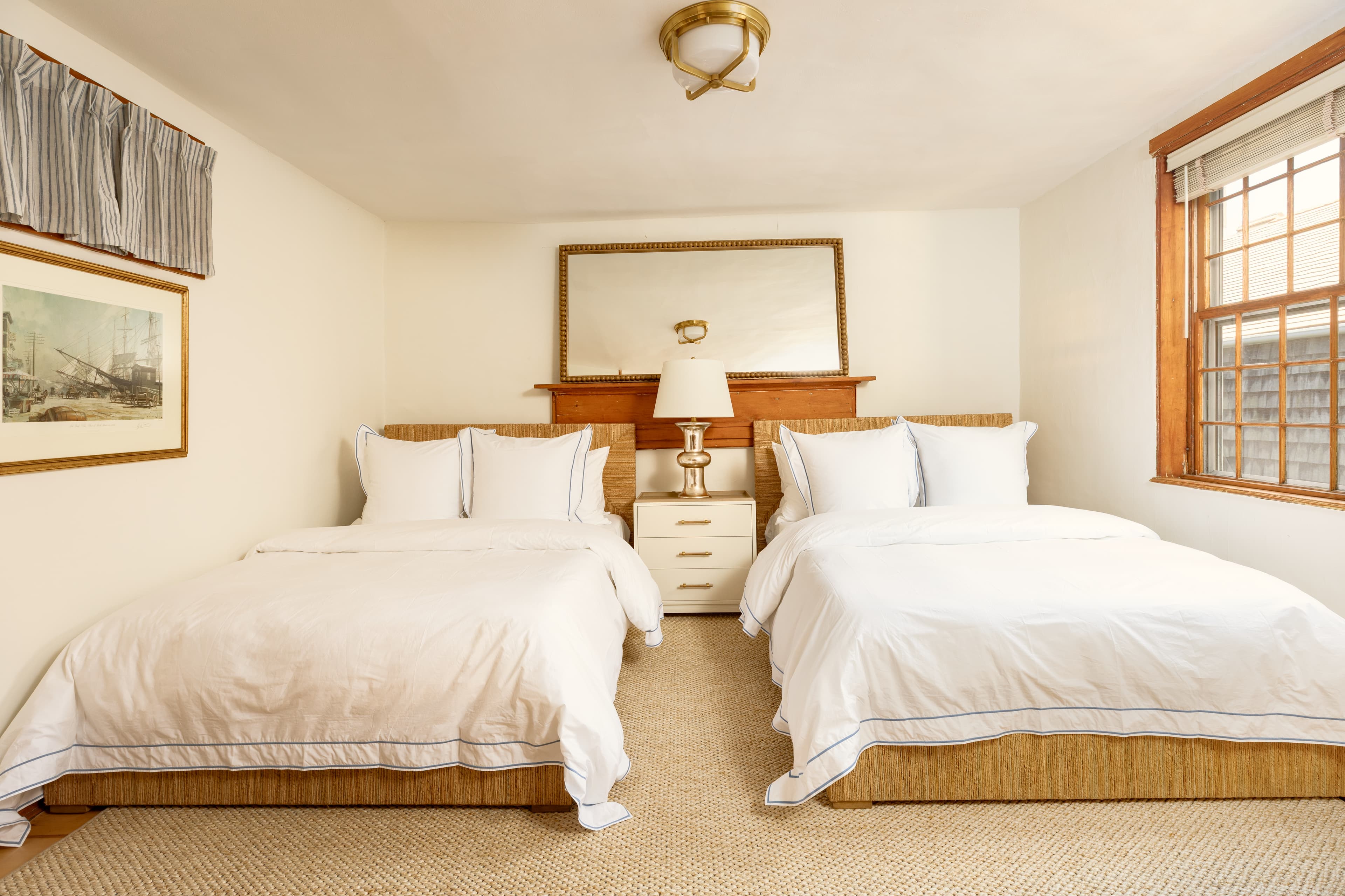 A comfortable bedroom featuring two beds with white linens, a nightstand with a lamp, a large mirror, and a window with wooden blinds.