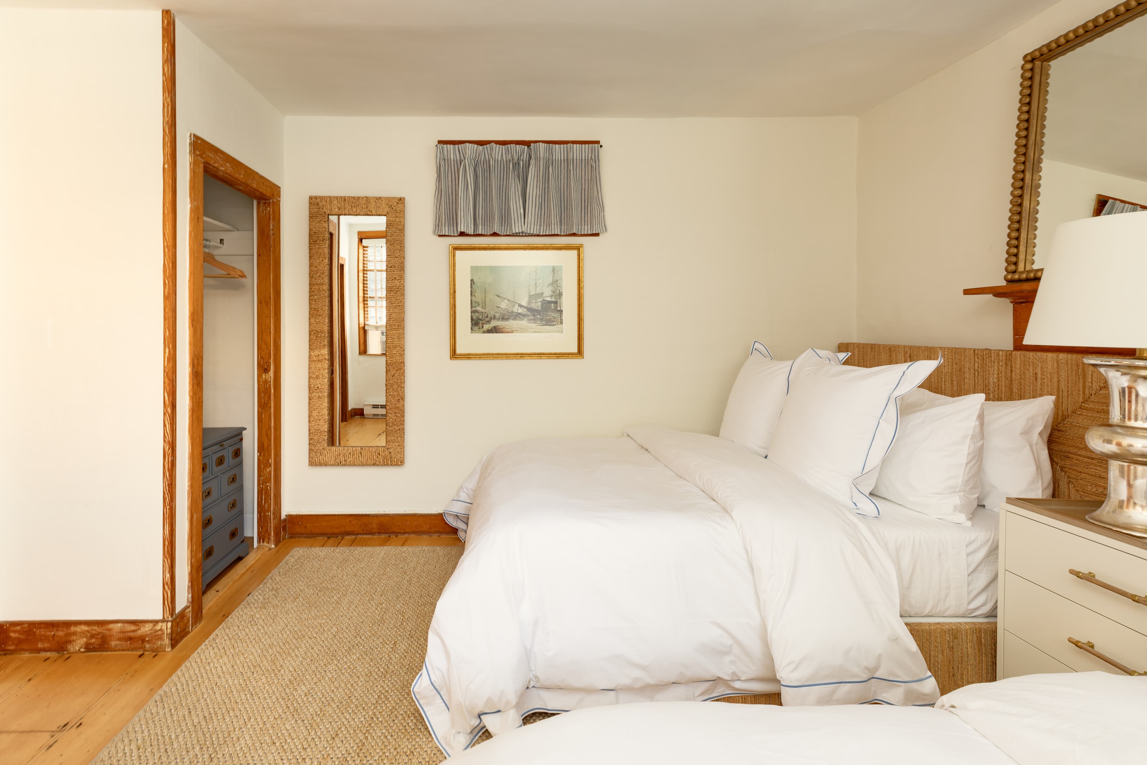 A cozy bedroom featuring two beds with white linens, warm wooden trim, a full-length mirror, and a framed picture above a low window.