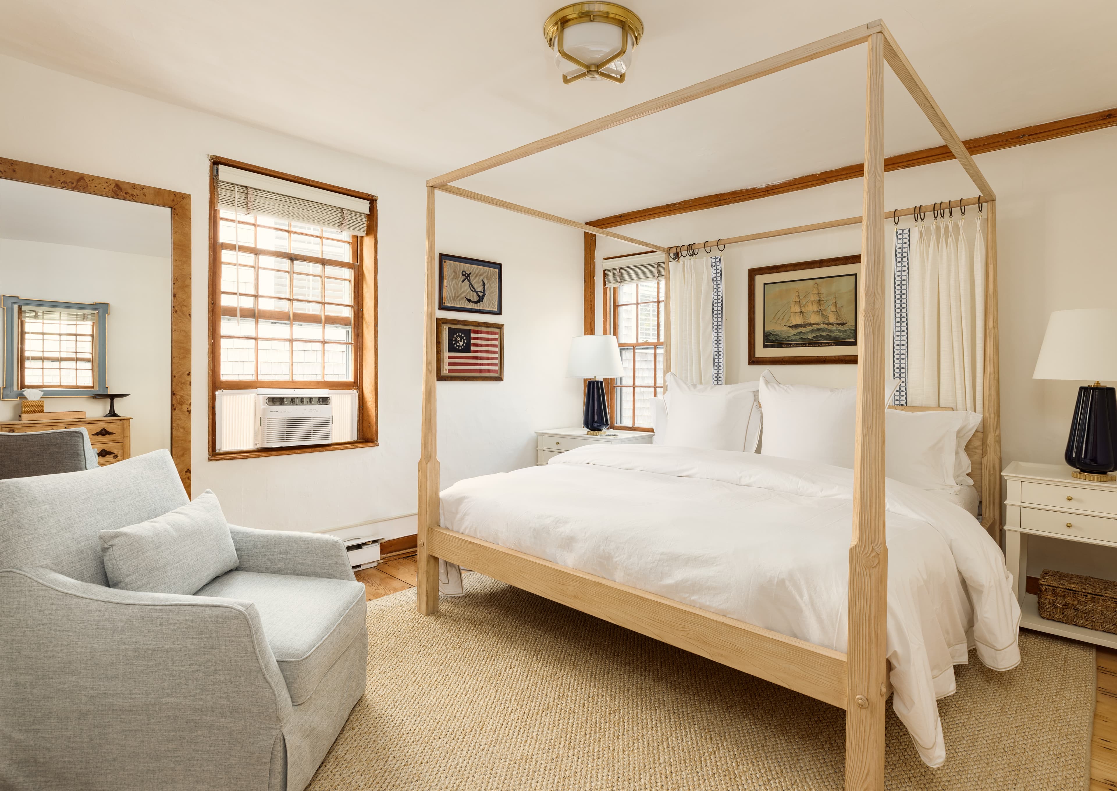 A serene bedroom with a light wood four-poster bed, crisp white linens, an armchair, and warm wooden accents, bathed in natural light.