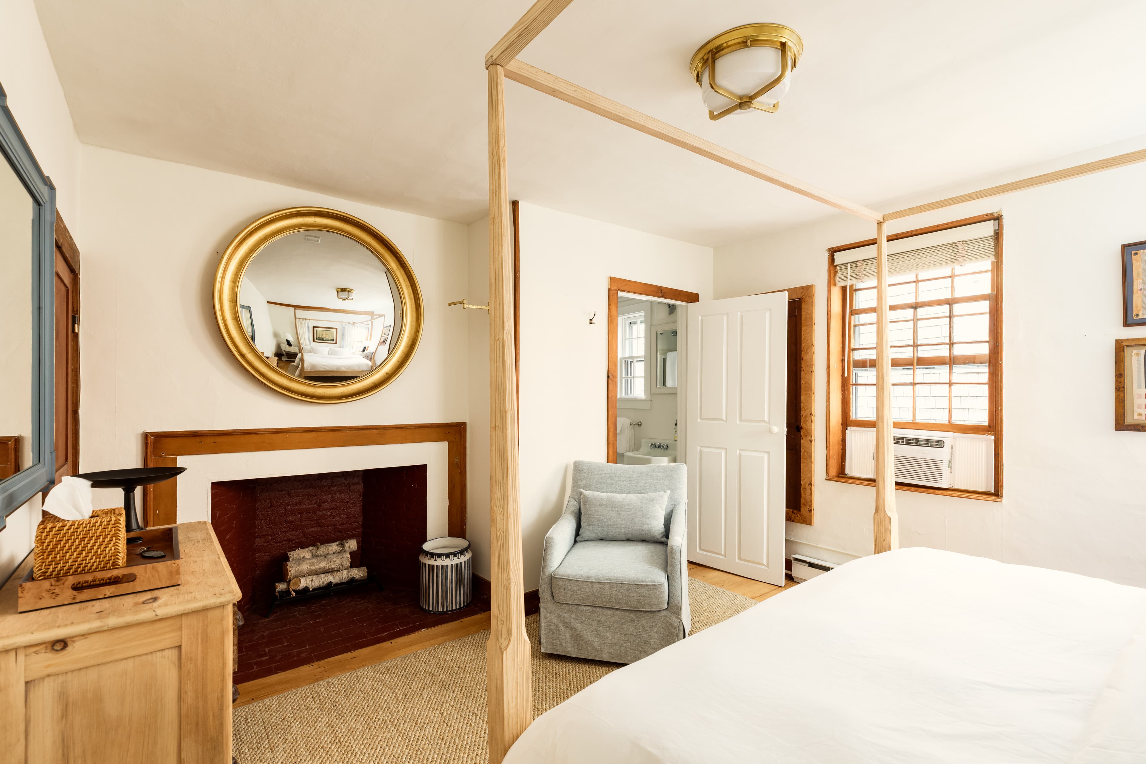 A cozy bedroom featuring a four-poster bed, a fireplace with a round mirror, an armchair, and wooden accents, creating a warm ambiance.