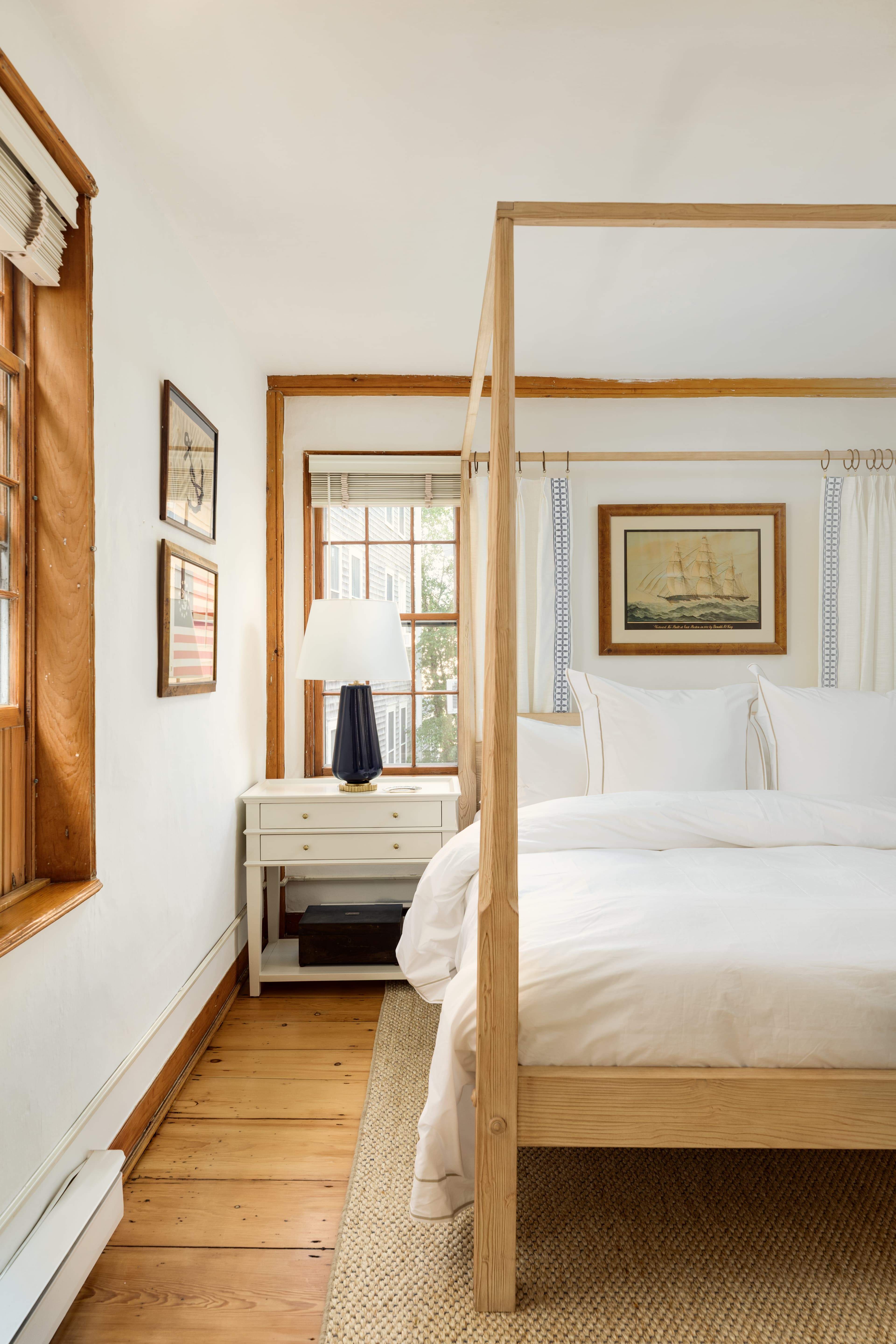 A cozy bedroom featuring a light wood four-poster bed with white linens, a nightstand with a lamp, and warm wooden window frames, bathed in natural light.