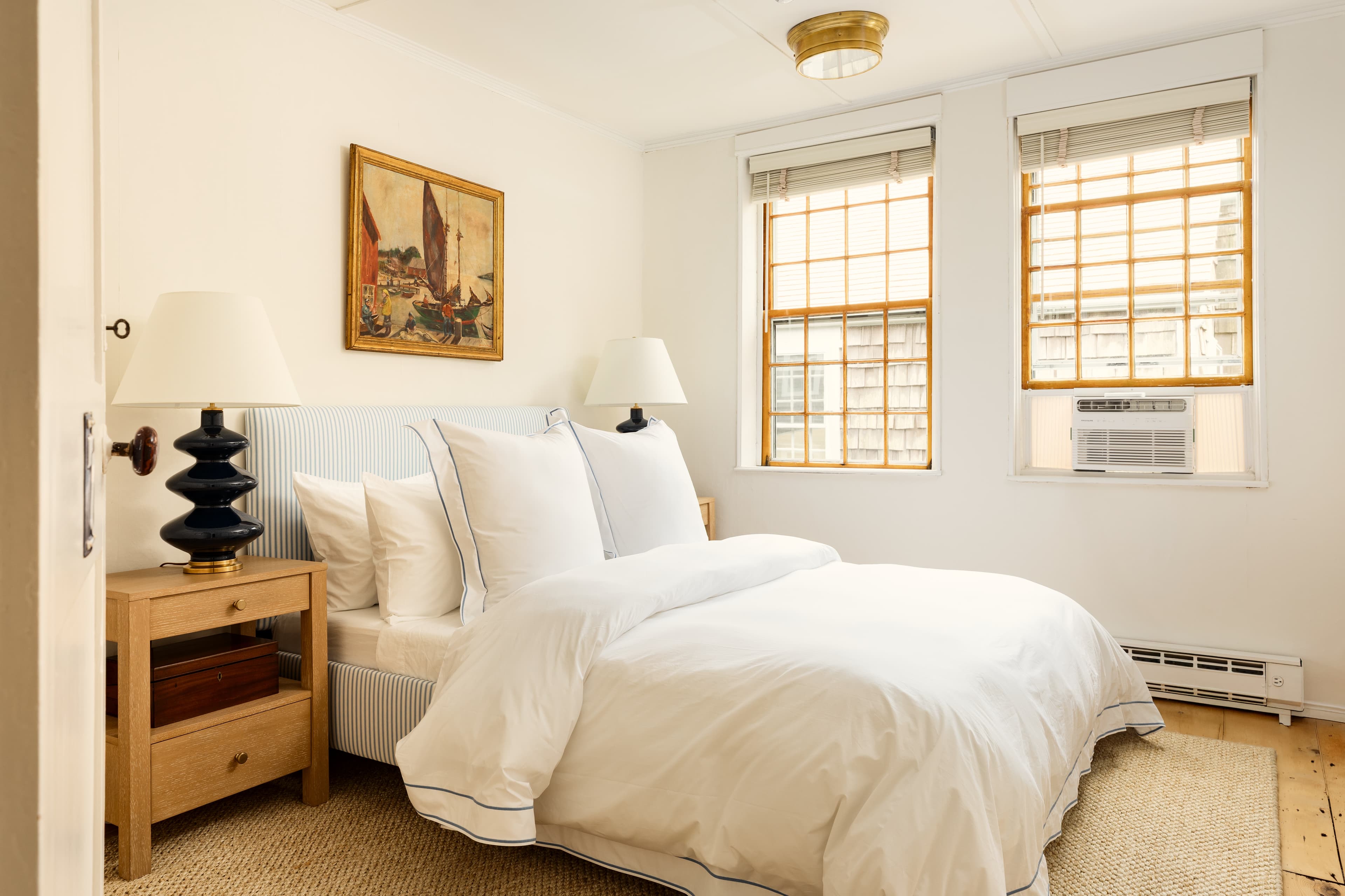 A serene bedroom featuring a neatly made bed with white linens, two windows with wooden blinds, and a nightstand with a dark lamp.
