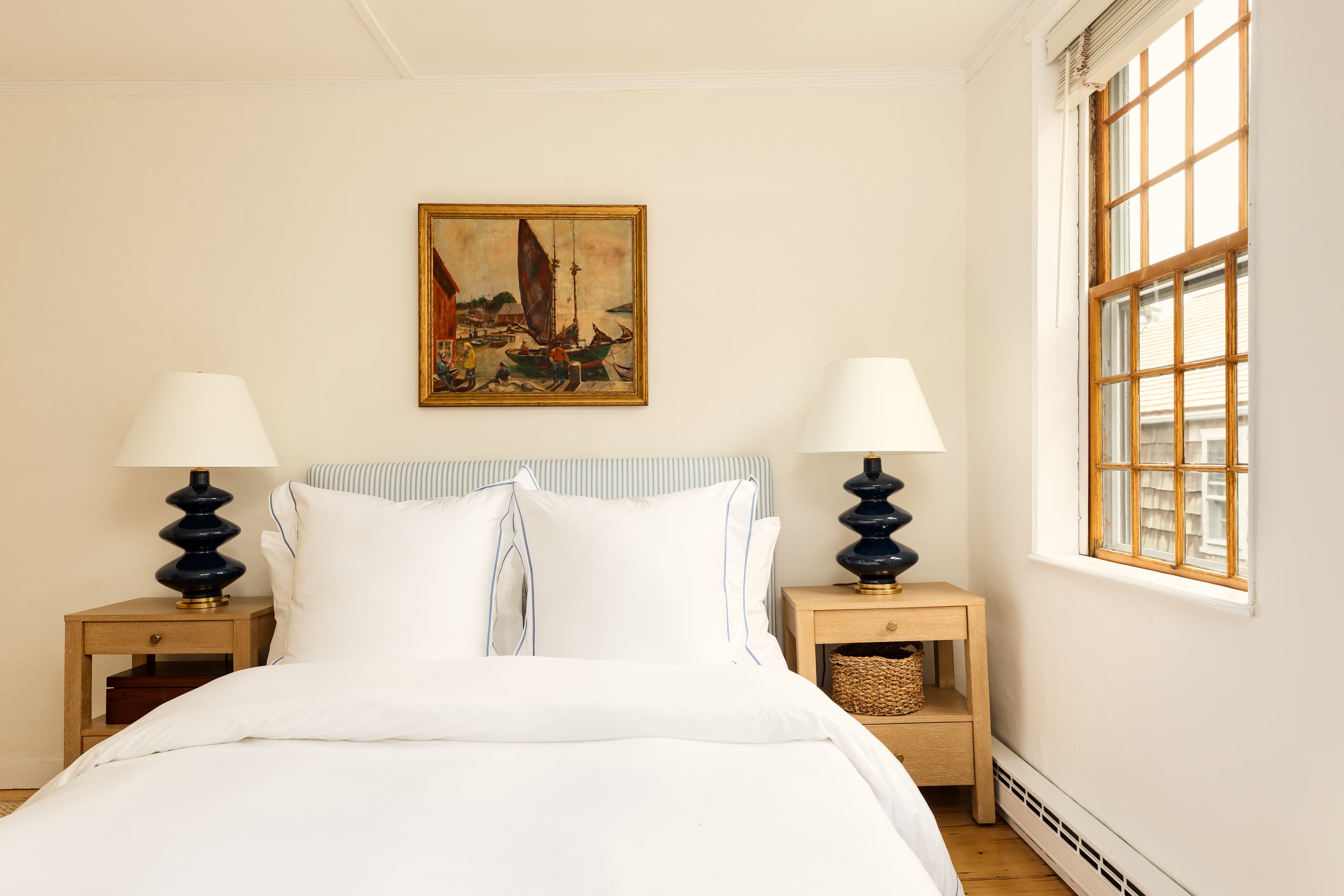 A neatly arranged bedroom featuring a bed with white linens, two wooden nightstands with blue lamps, and a window with wooden frames.