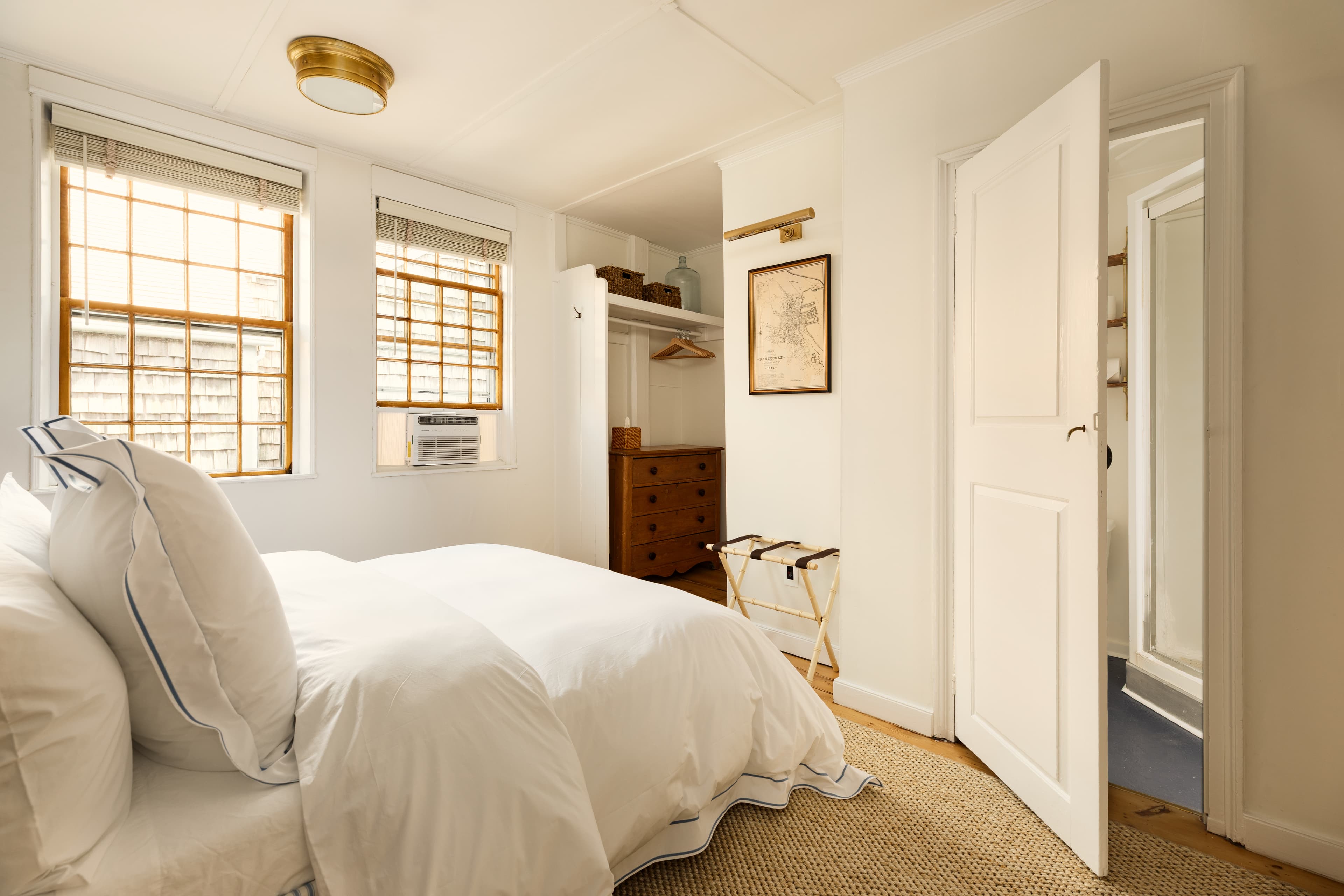 A bright bedroom featuring a white bed with ample pillows, two windows, an open closet, and a glimpse into another room.