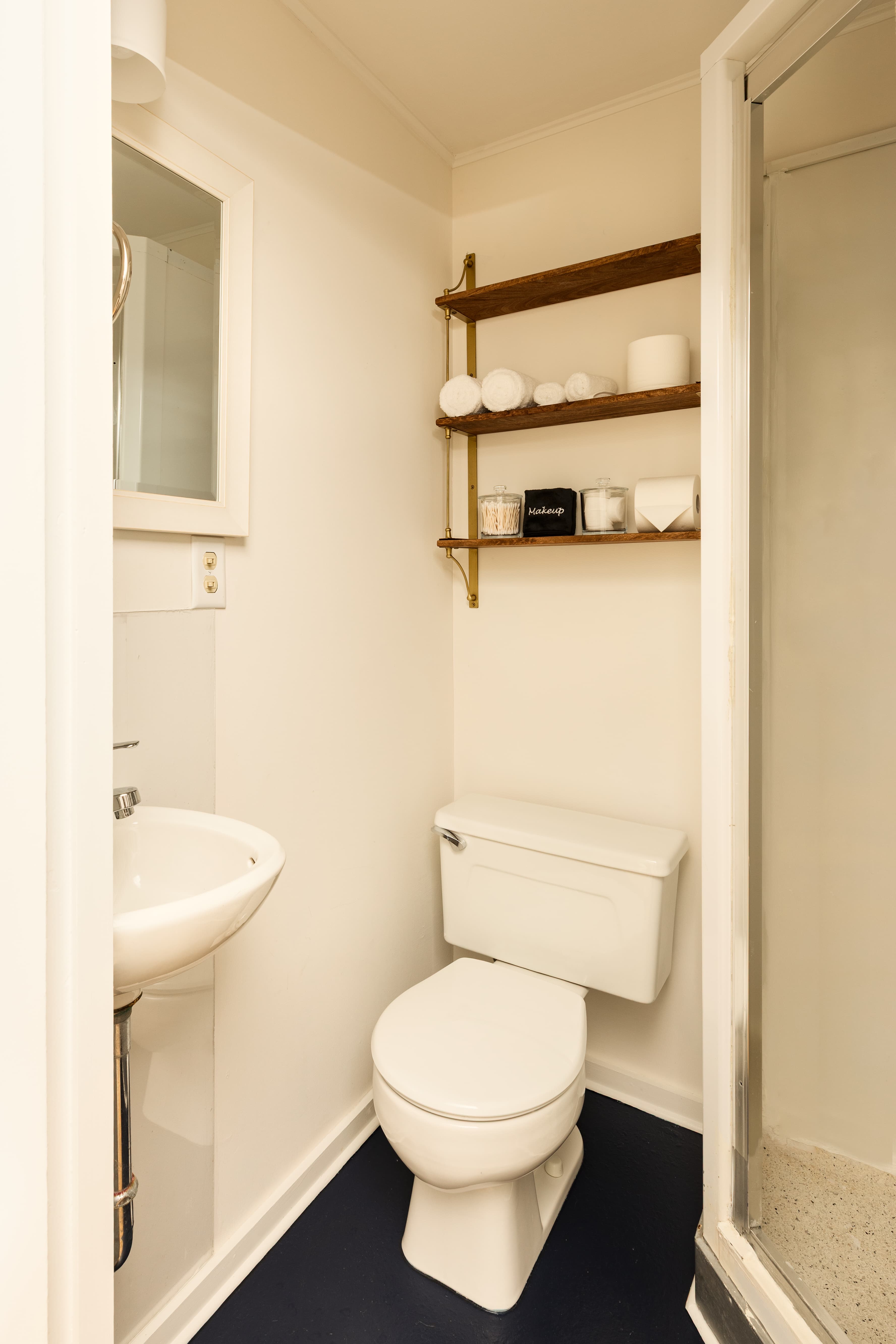 A compact, functional bathroom with a white toilet, pedestal sink, a glass-enclosed shower, and a wooden shelf with neatly stacked towels.