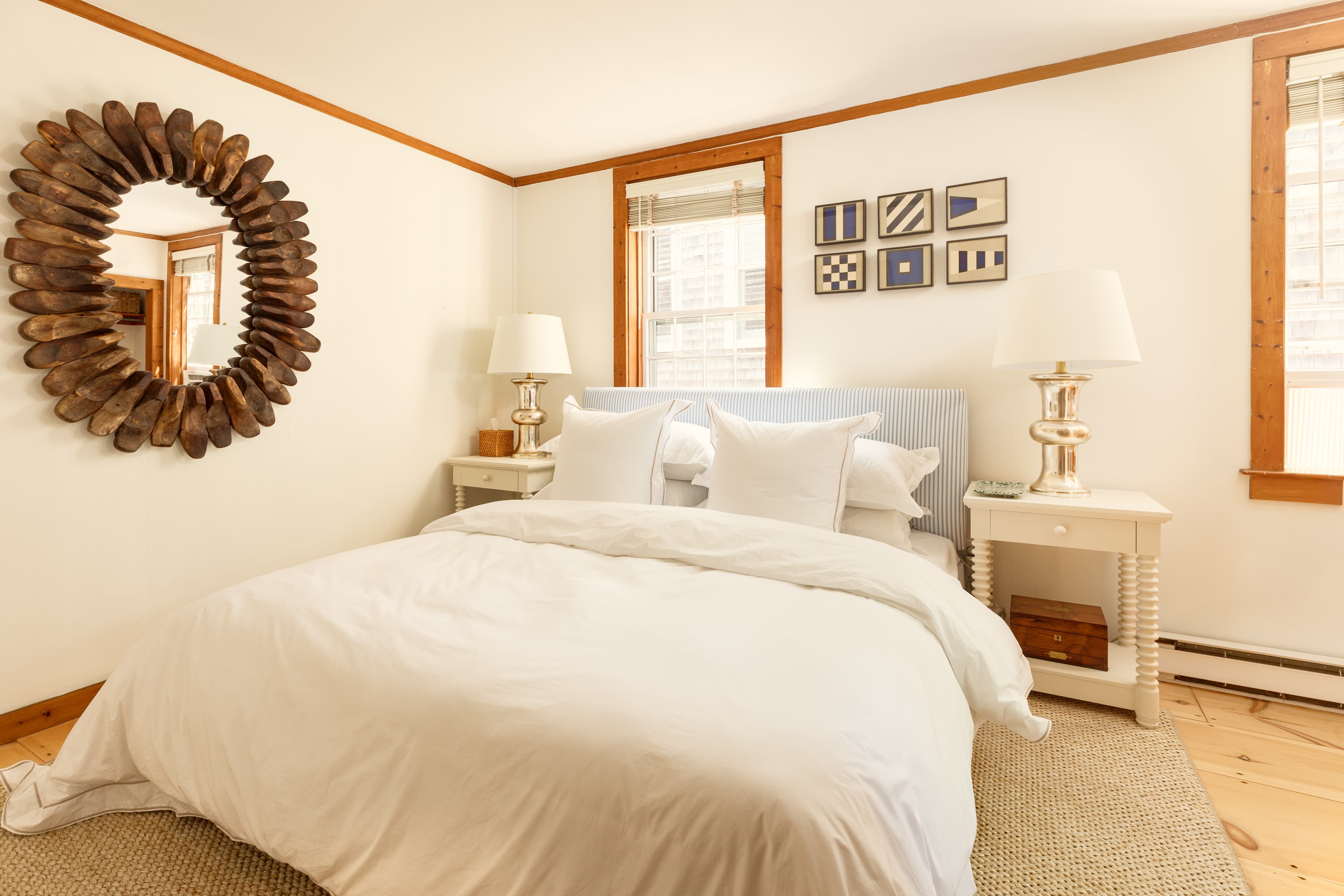 A bright, inviting bedroom featuring a white bed with light blue headboard, two windows, and nautical-themed decor, including a unique wooden mirror.