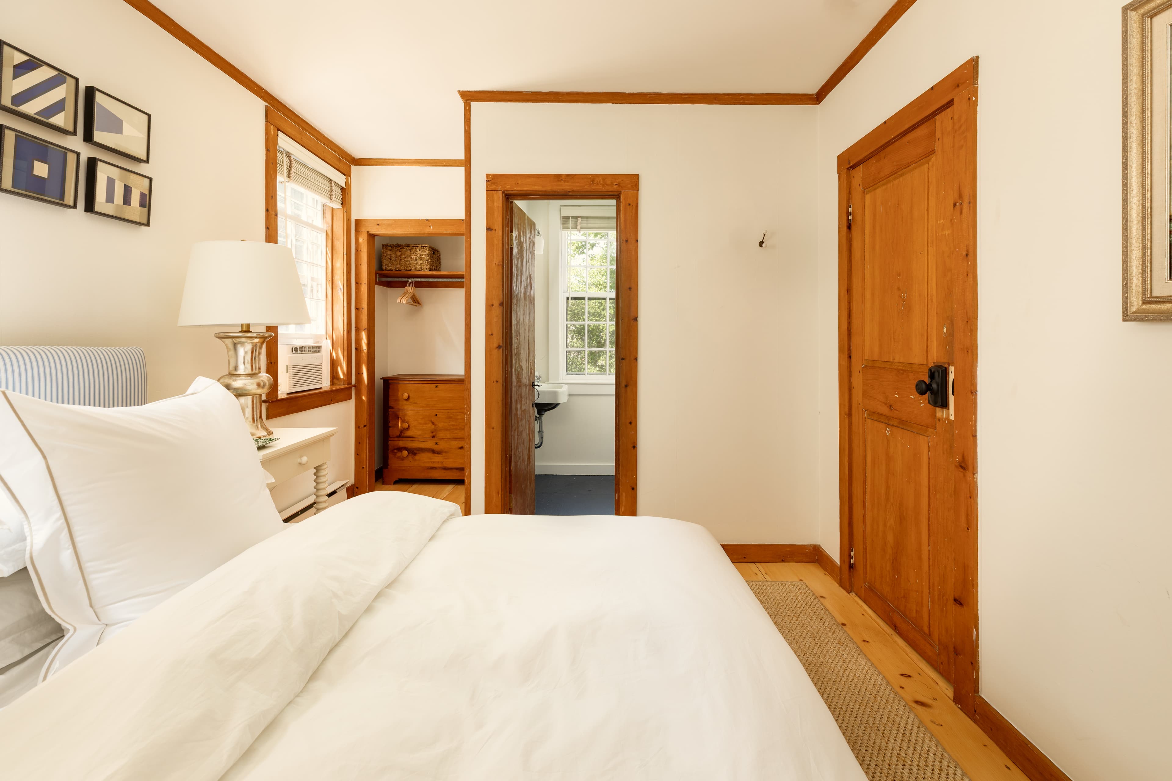 A serene bedroom featuring a white bed, warm wooden trim, and an open doorway leading to a bathroom. Nautical flags adorn the wall.