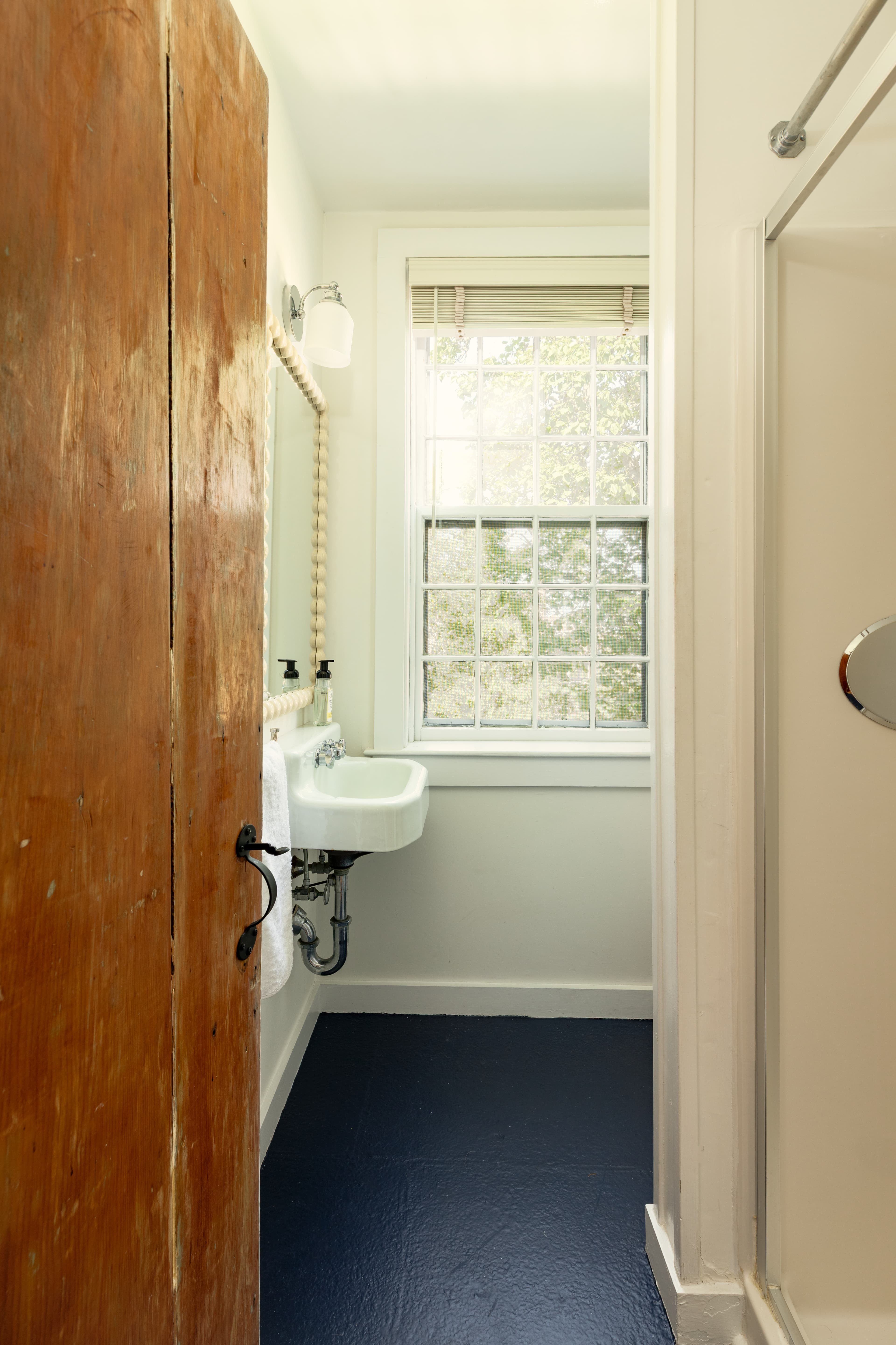 A clean, bright bathroom with a white pedestal sink under a window, a dark blue floor, and a glass-enclosed shower on the right.