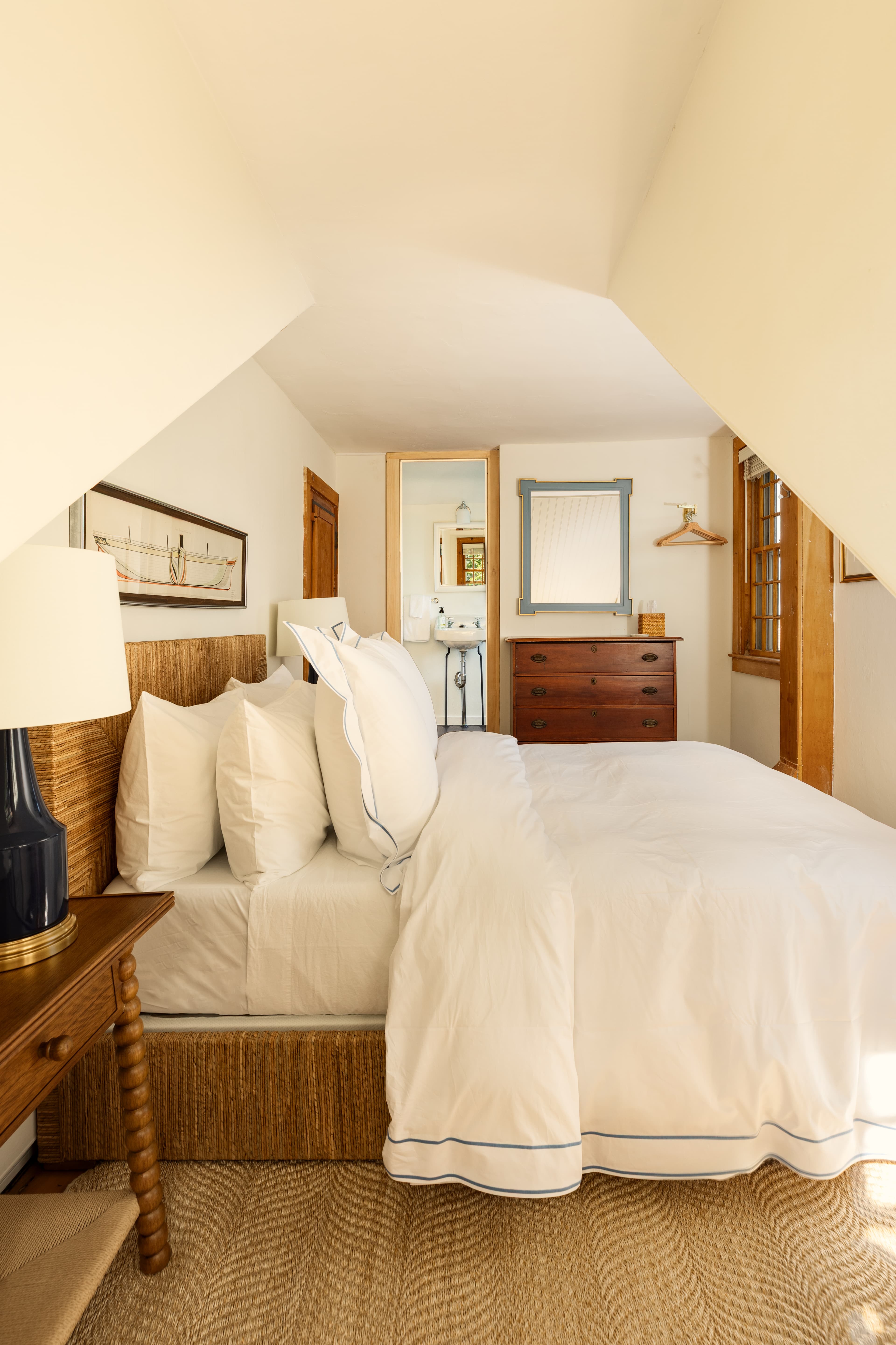 A bright attic bedroom with a bed featuring white linens and blue piping, a wooden nightstand with a dark lamp, and a dresser, bathed in sunlight.