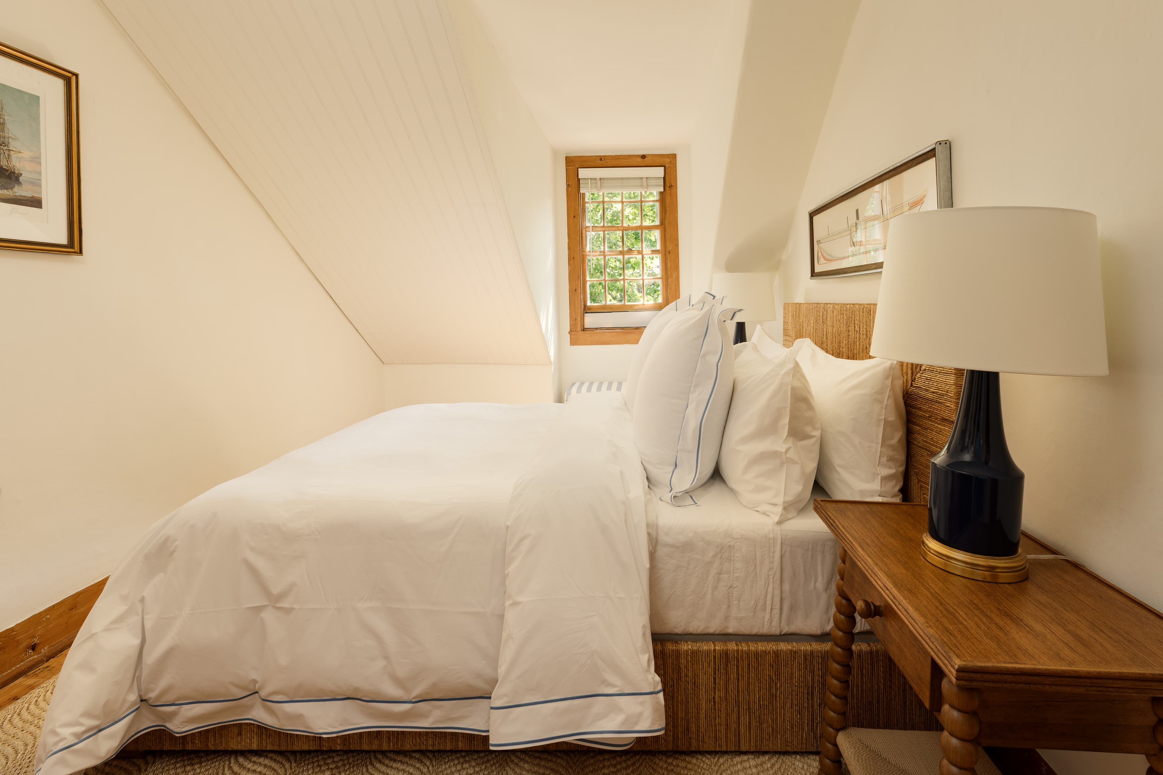 A cozy attic bedroom featuring a bed with white linens and blue piping, a wooden nightstand with a dark lamp, and a window offering natural light.