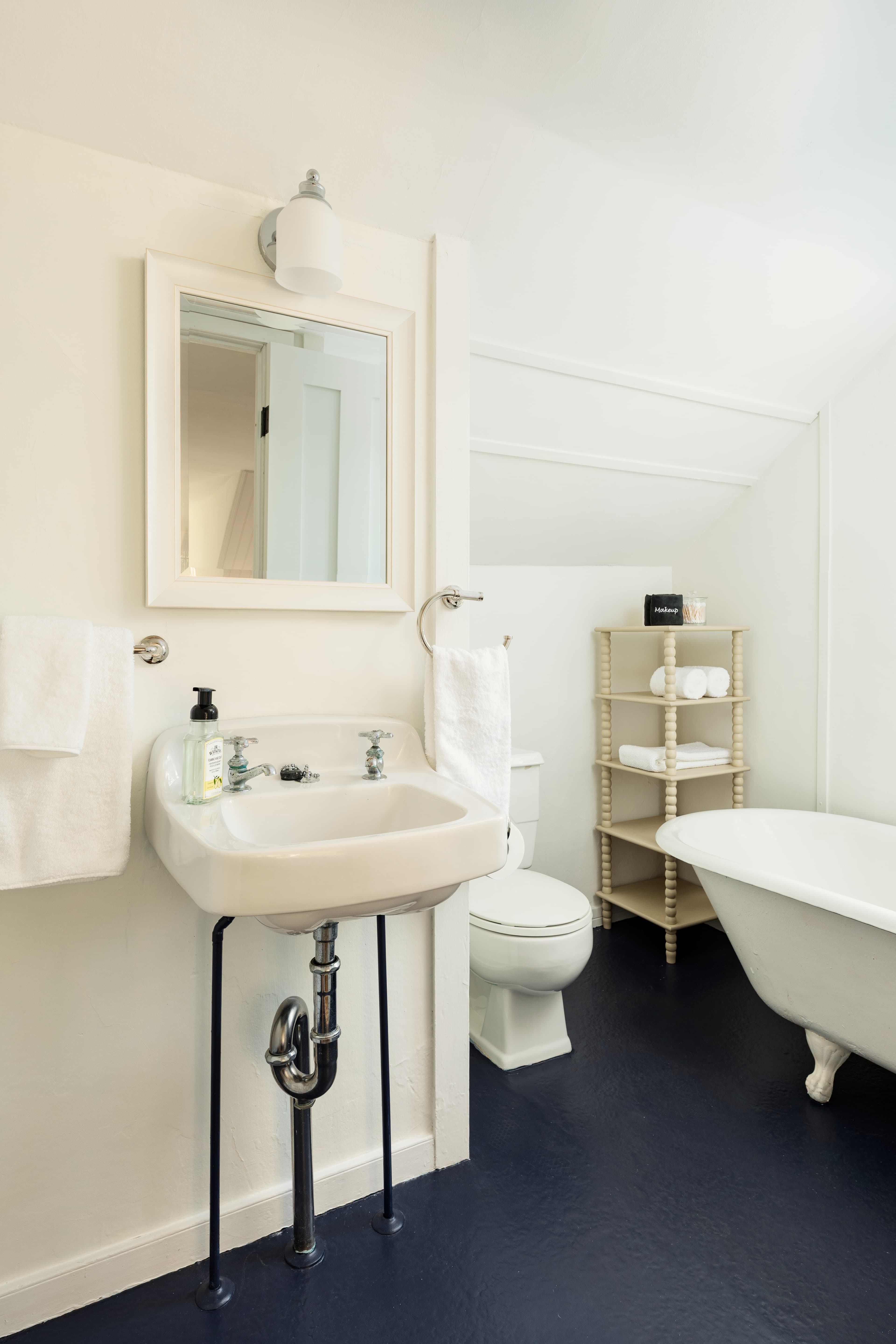 A charming bathroom featuring a white pedestal sink, toilet, and claw-foot tub, with a dark blue floor and a light-framed mirror.