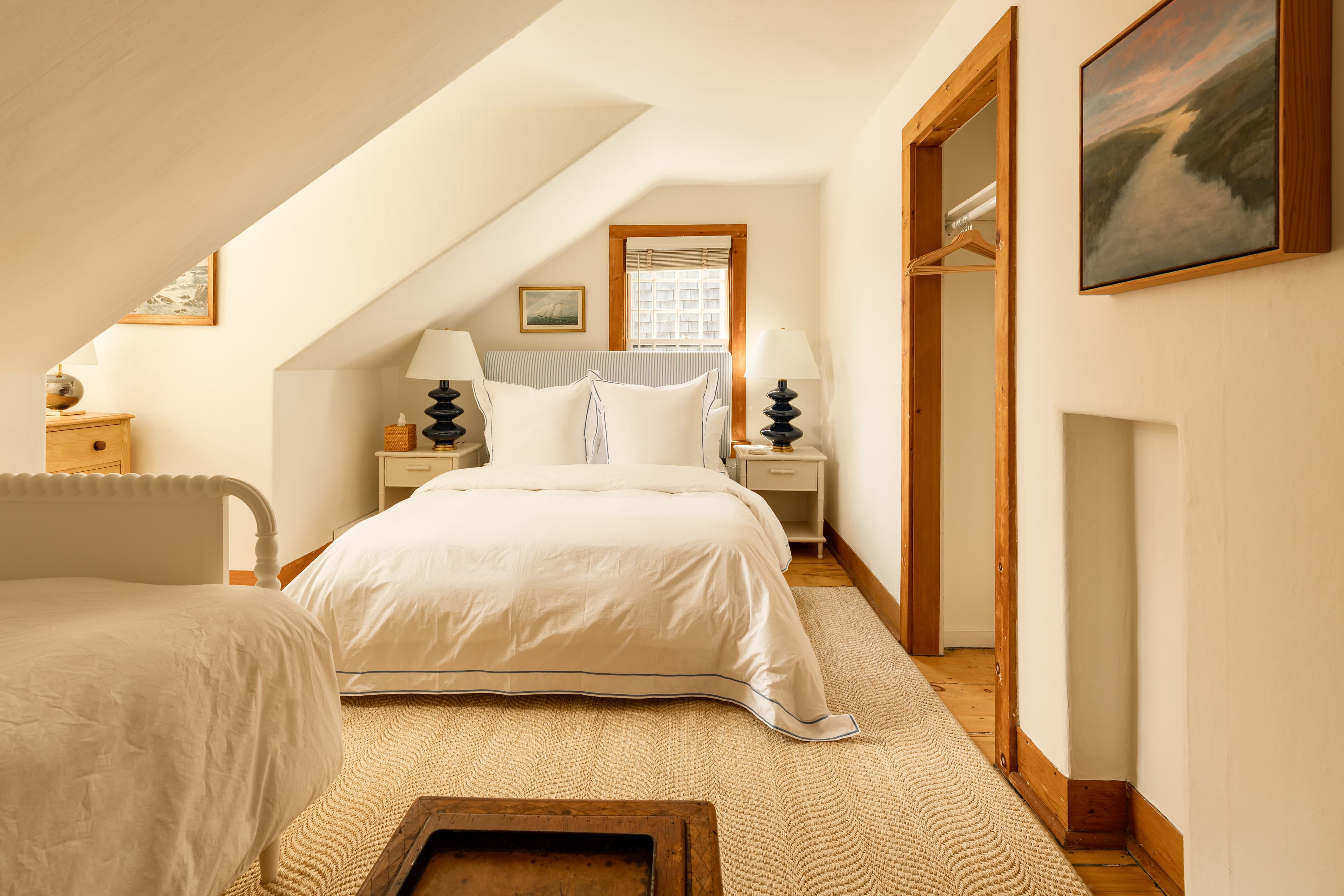 A spacious attic bedroom featuring a large bed with white linens, a smaller bed, and warm wooden accents, all bathed in natural light.