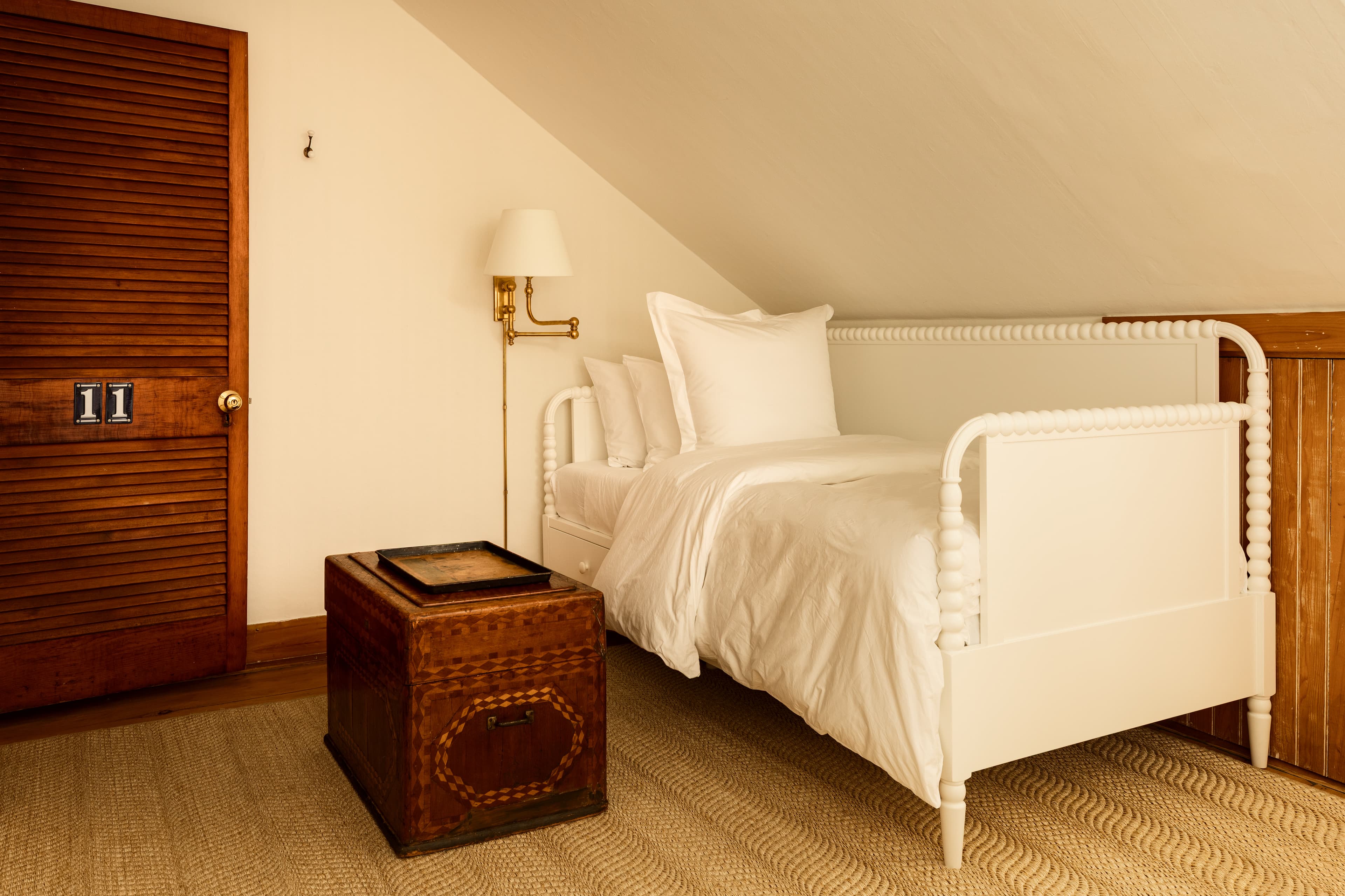 A cozy attic room featuring a white single bed, a unique wooden chest, and a golden wall lamp, with a wooden door adding warmth.