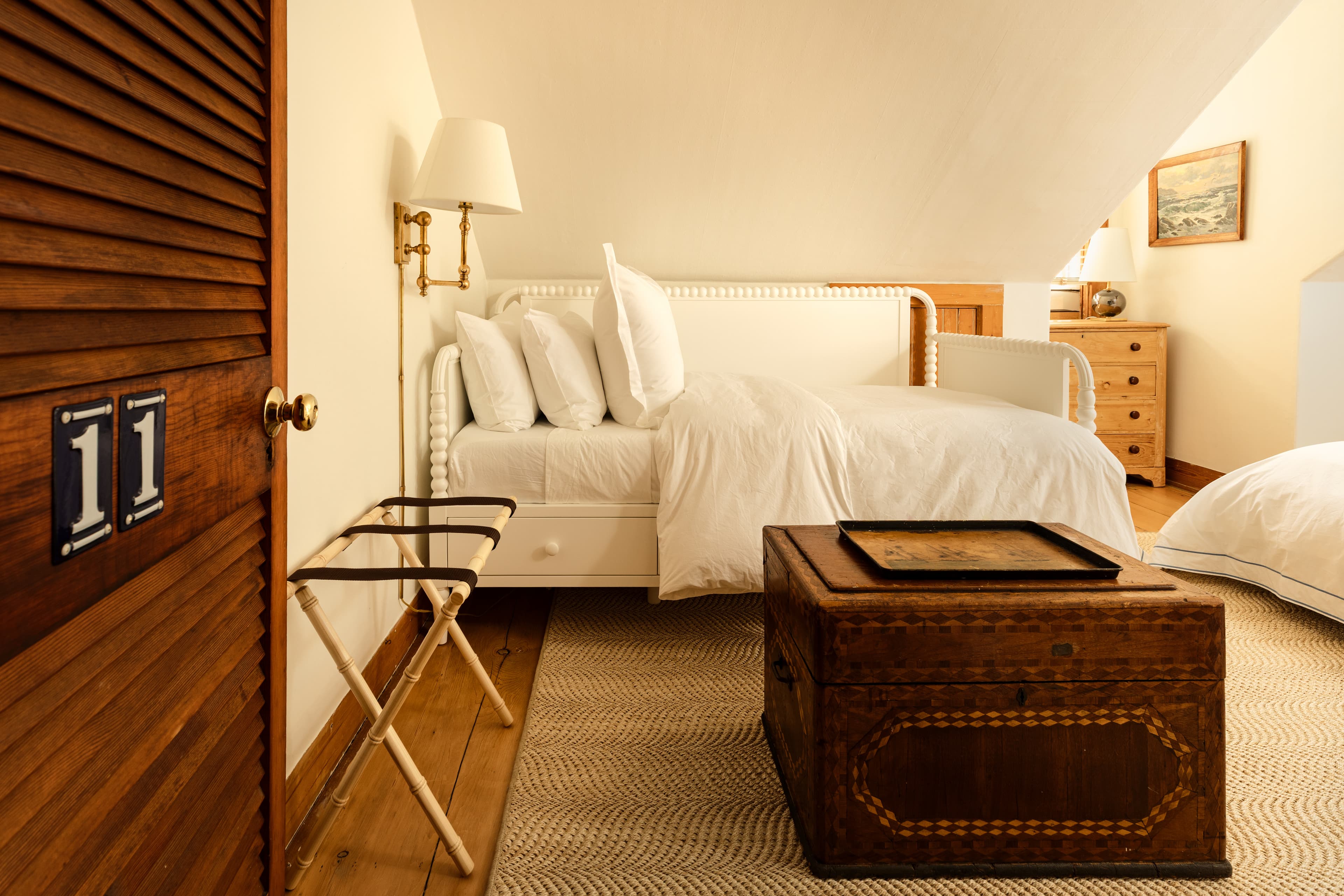 A charming attic bedroom with two beds, a unique wooden chest, a luggage rack, and a numbered door, all bathed in soft light.