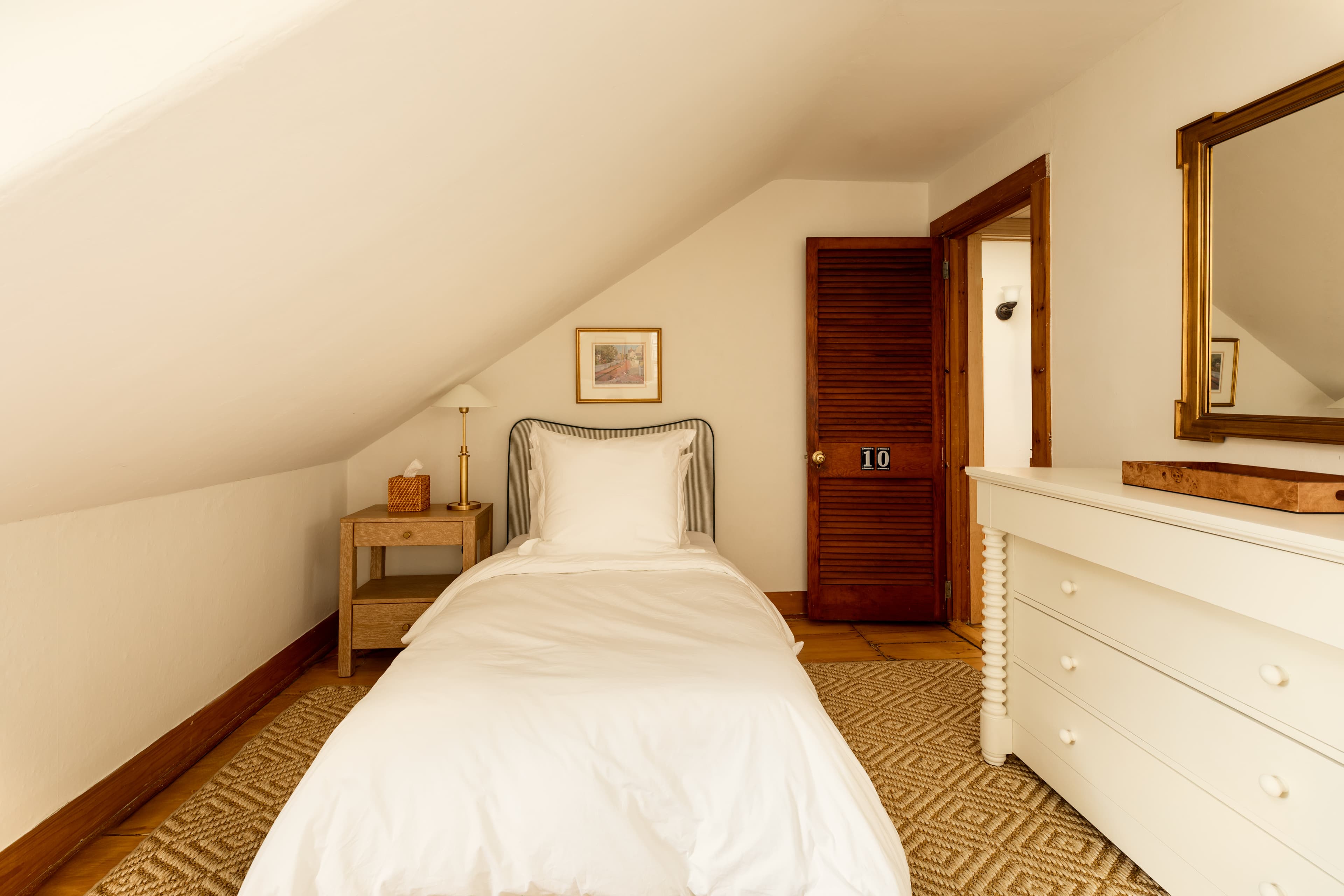 A serene attic bedroom featuring a single bed with white linens, a wooden nightstand, a white dresser, and a wooden door.