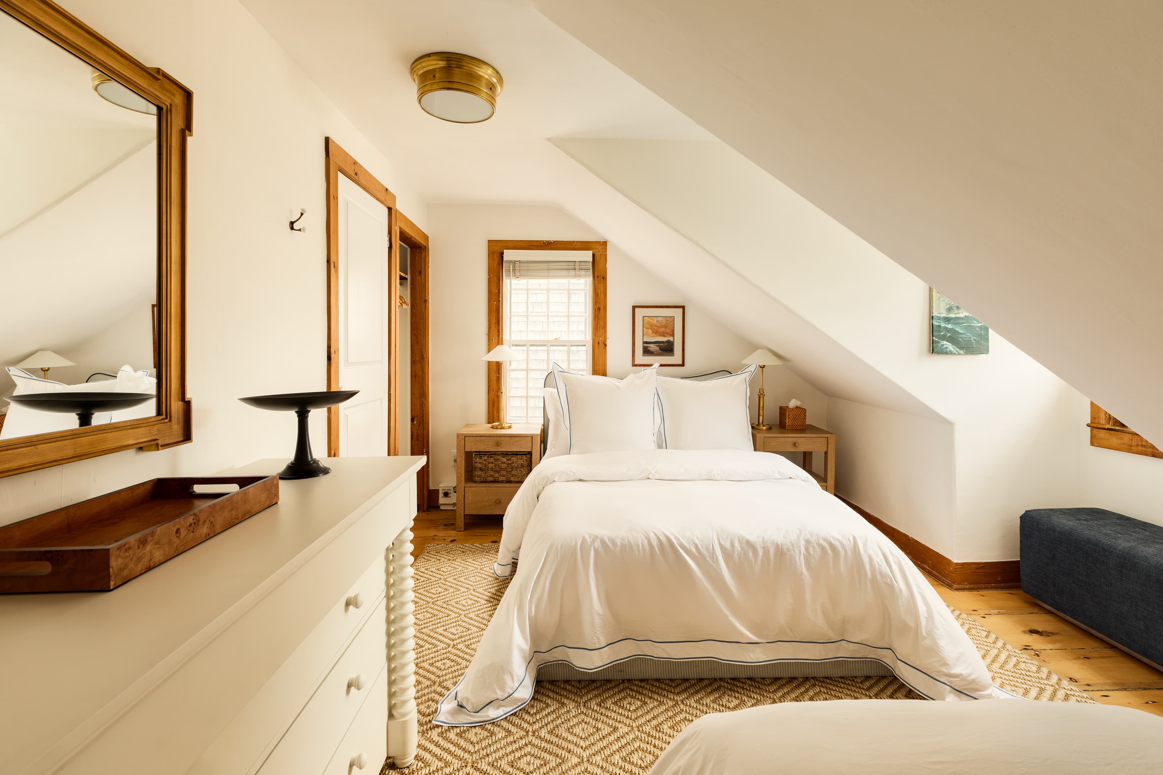 A spacious attic bedroom featuring a white bed with crisp linens, a white dresser with a mirror, and warm wooden accents, bathed in natural light.