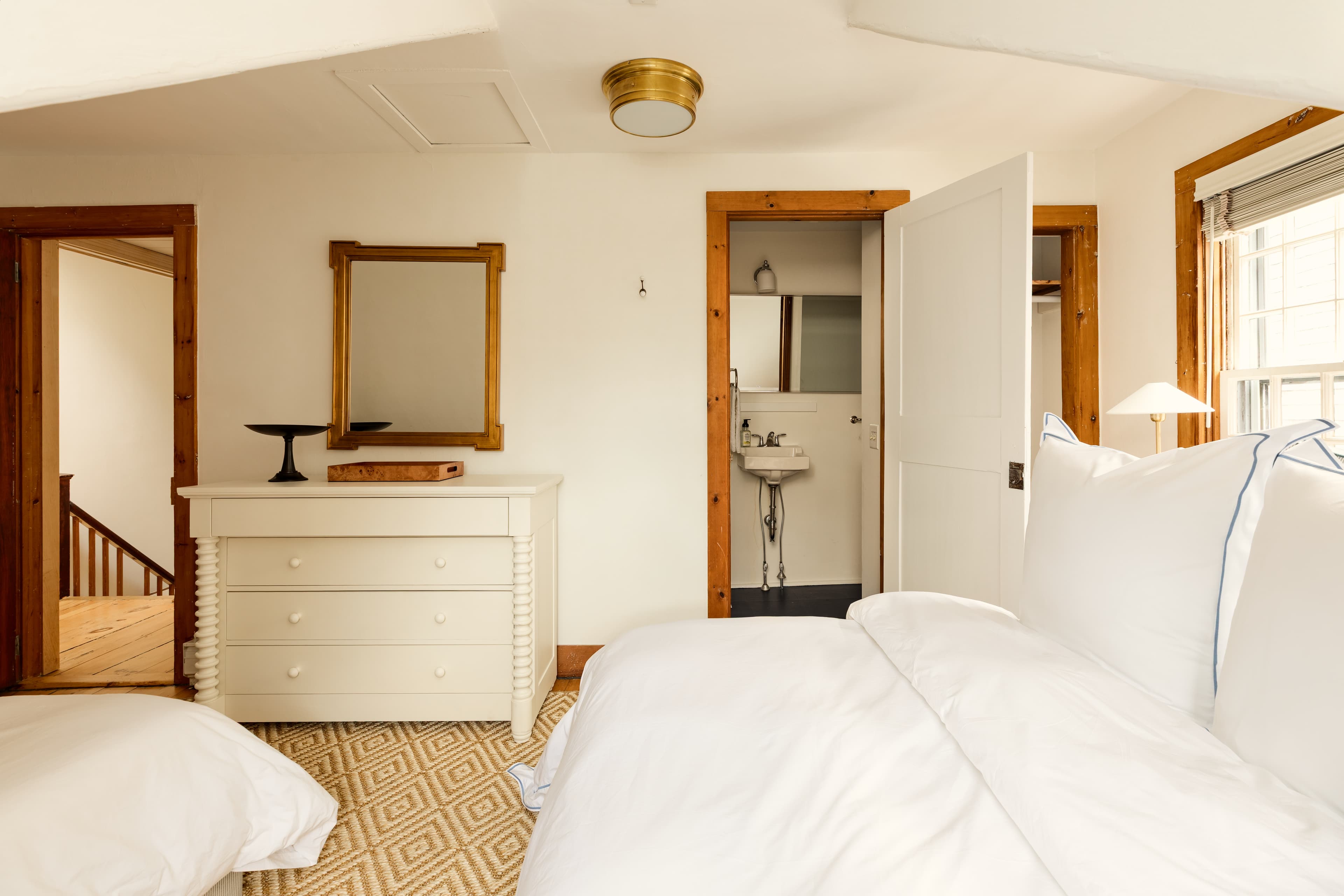 A bright attic bedroom with two white beds, a dresser with a mirror, and an open door revealing a bathroom with a sink.