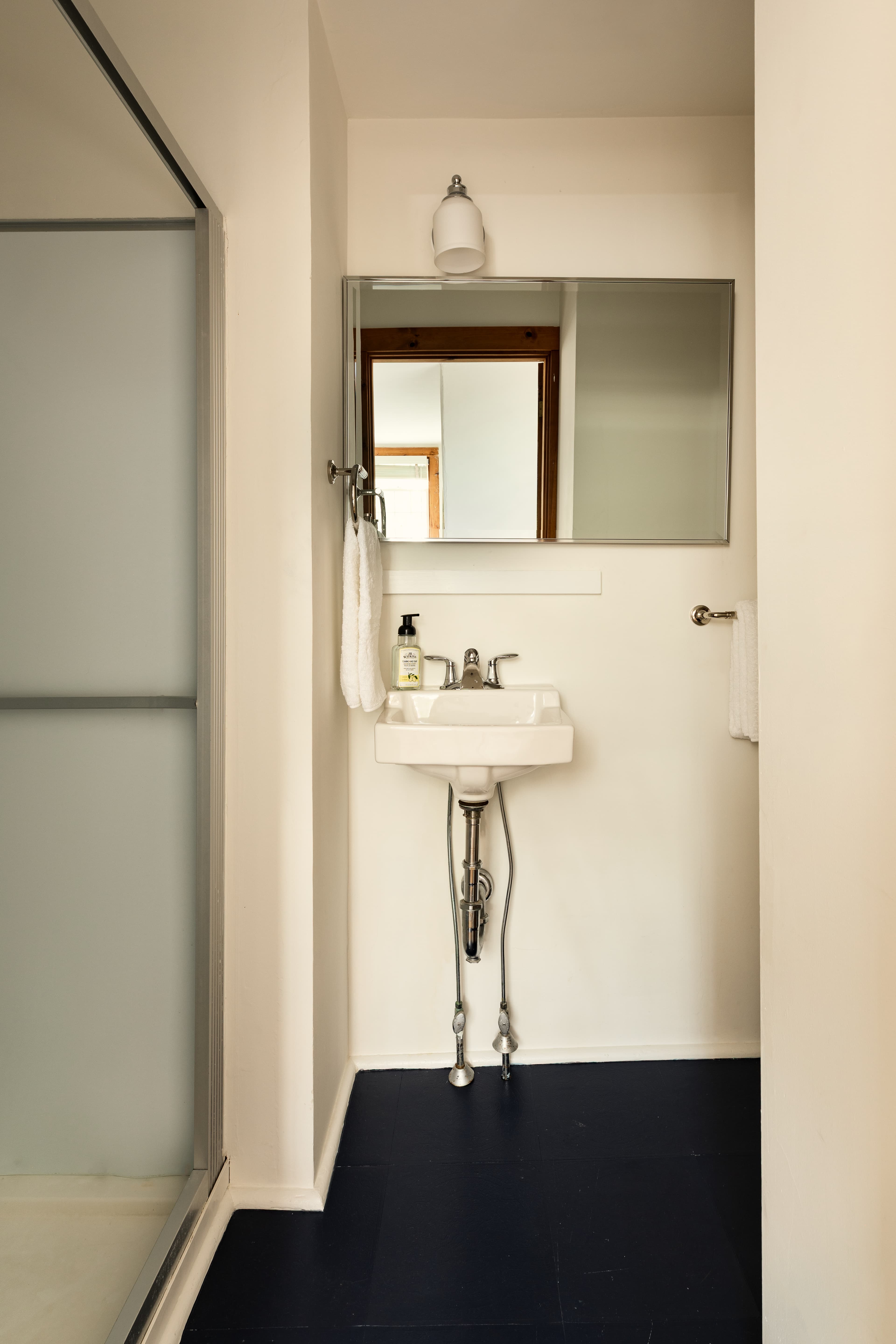 A clean, compact bathroom with a white pedestal sink, a mirror, and a frosted glass shower door, all on a dark blue floor.
