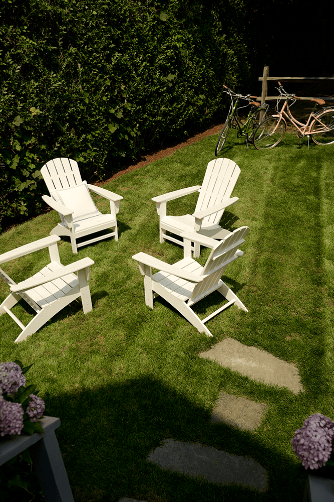 Four white Adirondack chairs placed in a circle, on a green lawn, with a hedgerow along one side and bicycles leaning against a wooden fence along the other side.