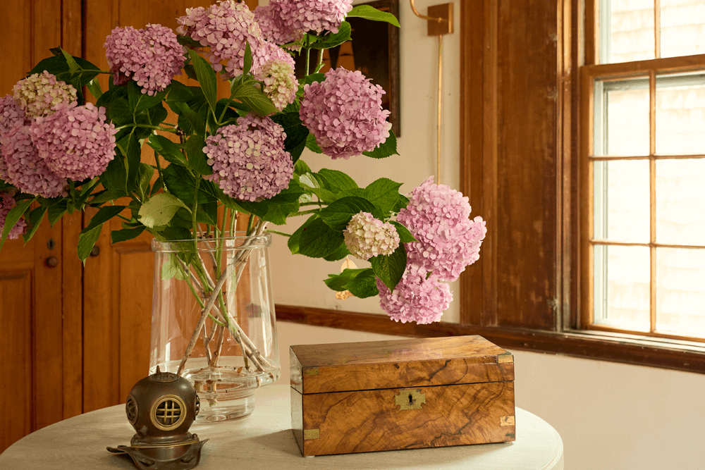 A vase of pink hydrangeas beside a wooden box and a decorative diving helmet on a table near a window.